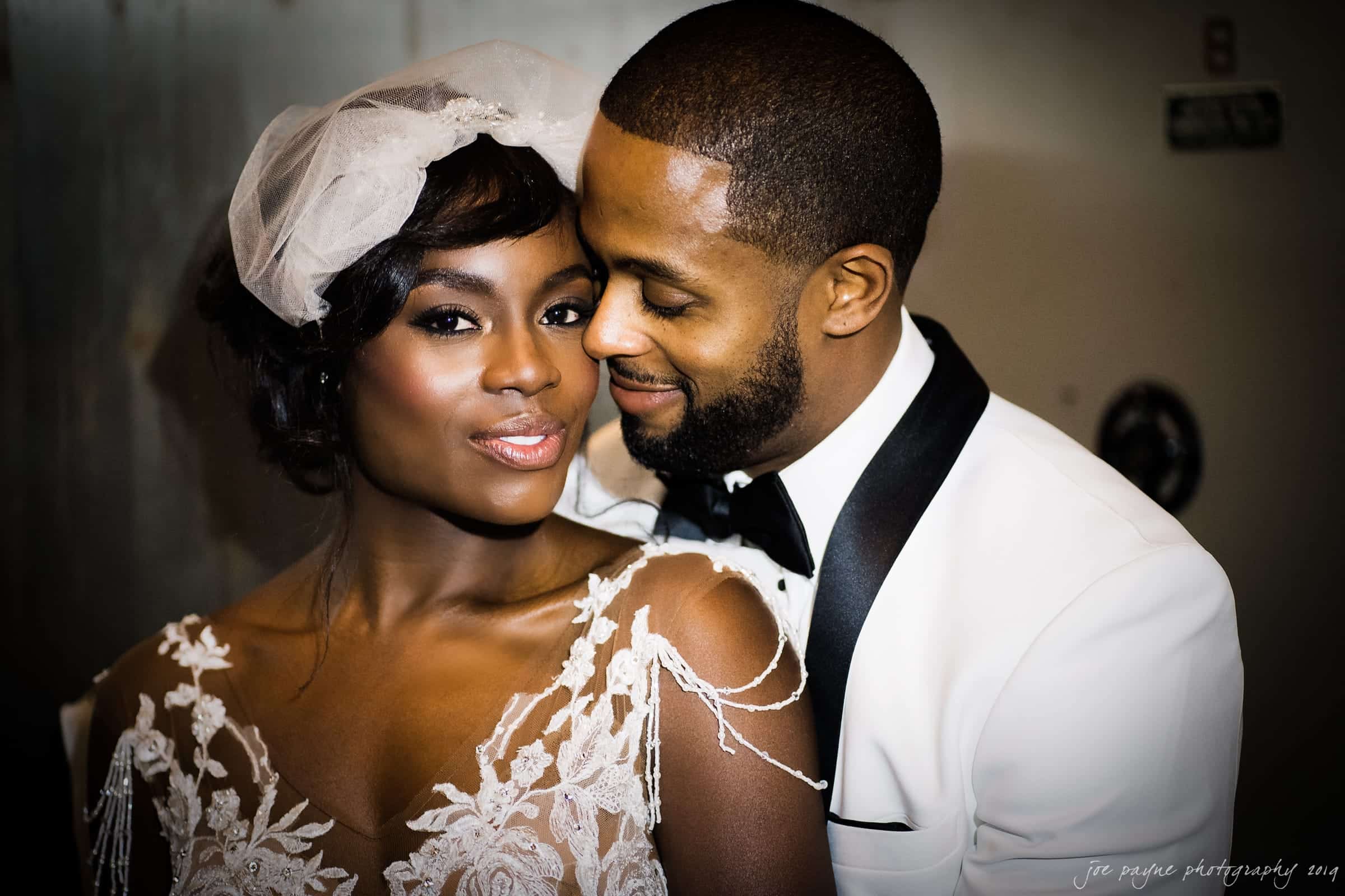 21c museum hotel durham wedding - kortne & terry A bride in an elegant white lace wedding dress and veil stands with her husband, who is wearing a white tuxedo. They are close together, sharing a tender, joyful moment, with the groom smiling affectionately at the bride.