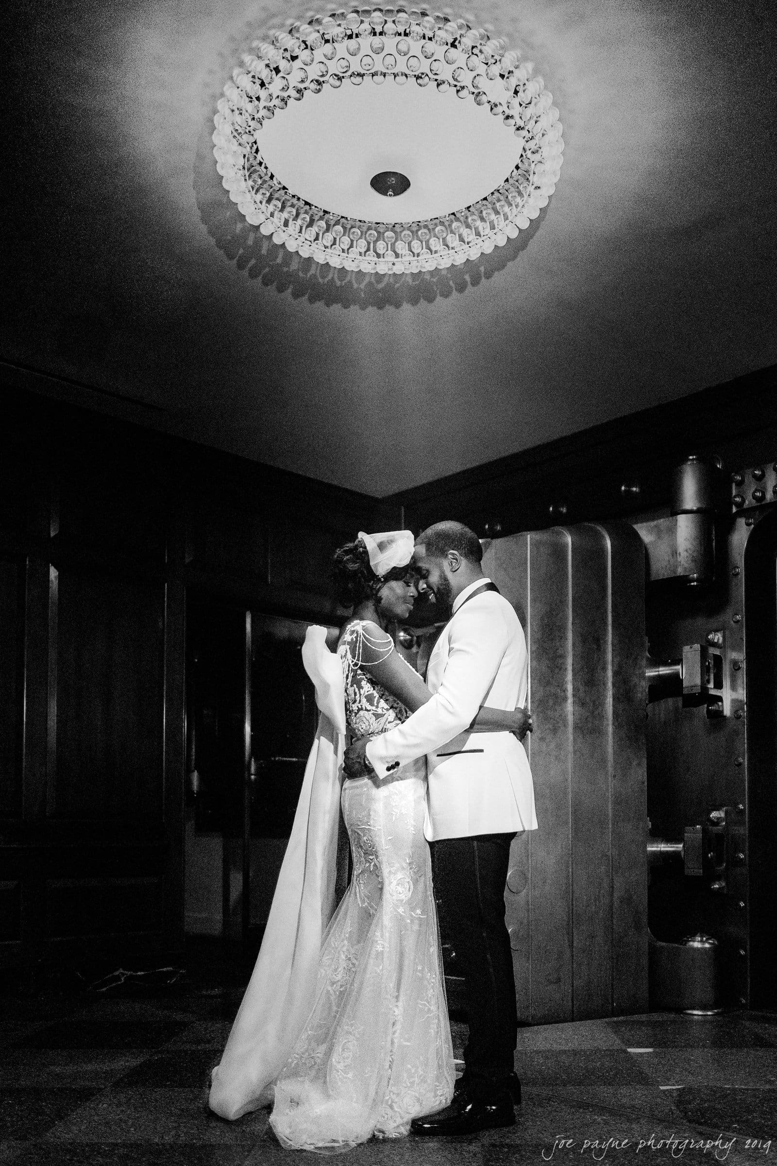 21c museum hotel durham wedding - kortne & terry A couple embraces closely in an elegant room, with an ornate chandelier overhead. The bride wears a flowing dress and veil, and the groom is in a white tuxedo jacket. The atmosphere is intimate and romantic.