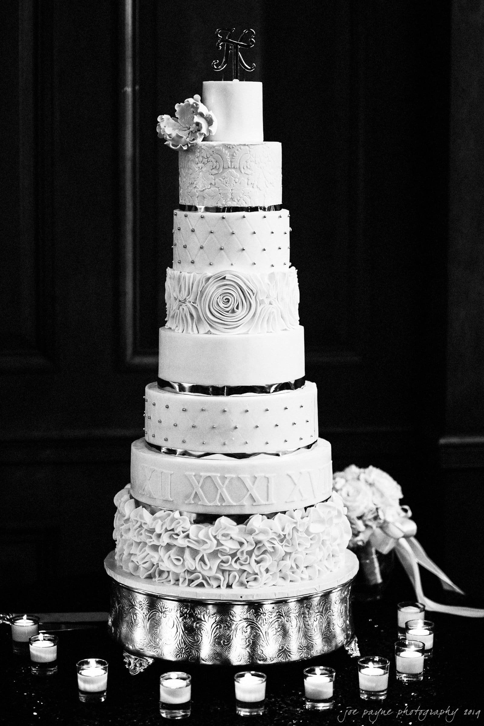 21c museum hotel durham wedding - kortne & terry A tall, ornate wedding cake with multiple tiers, each adorned with unique patterns and textures, topped with an initial monogram. The cake sits on a decorated stand surrounded by small, lit candles.