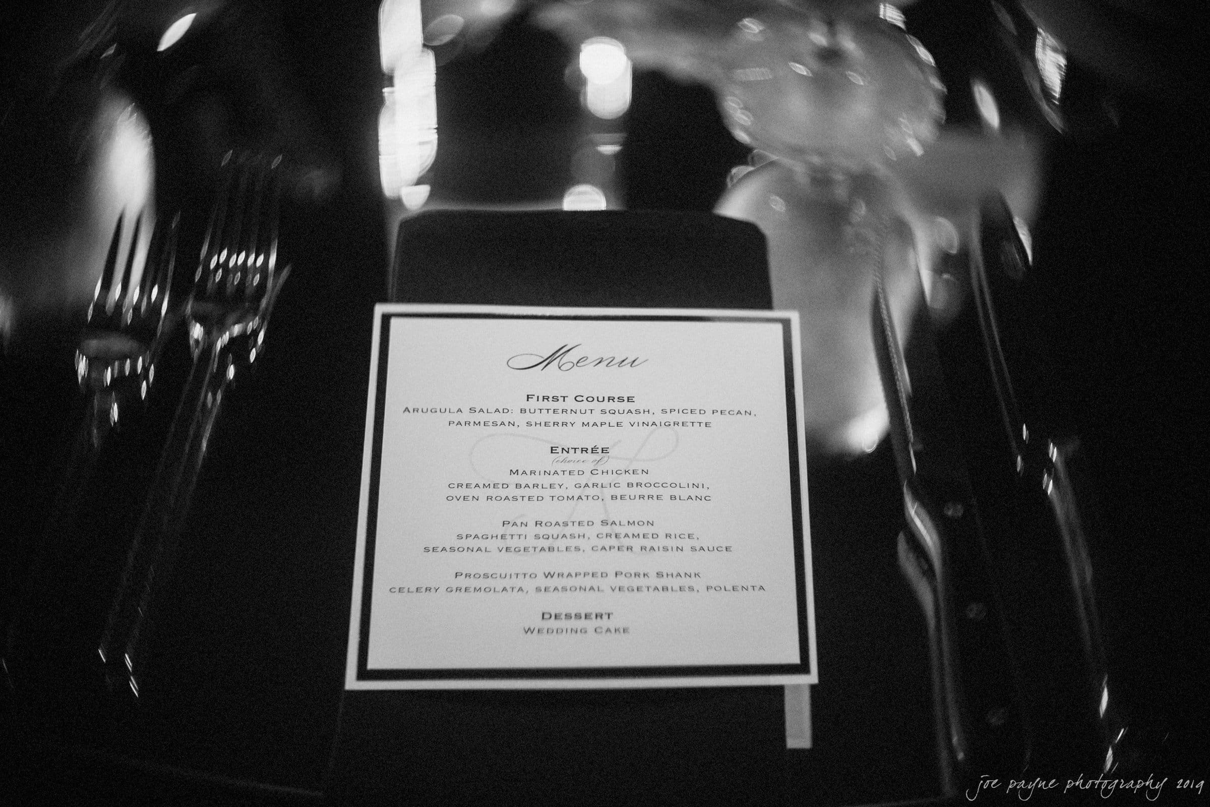 21c museum hotel durham wedding - kortne & terry A black and white image of a wedding menu on a dark tablecloth. The menu lists courses such as arugula salad, marinated chicken, pan-roasted salmon, prosciutto-wrapped pork shank, and wedding cake. Elegant place settings surround the menu.