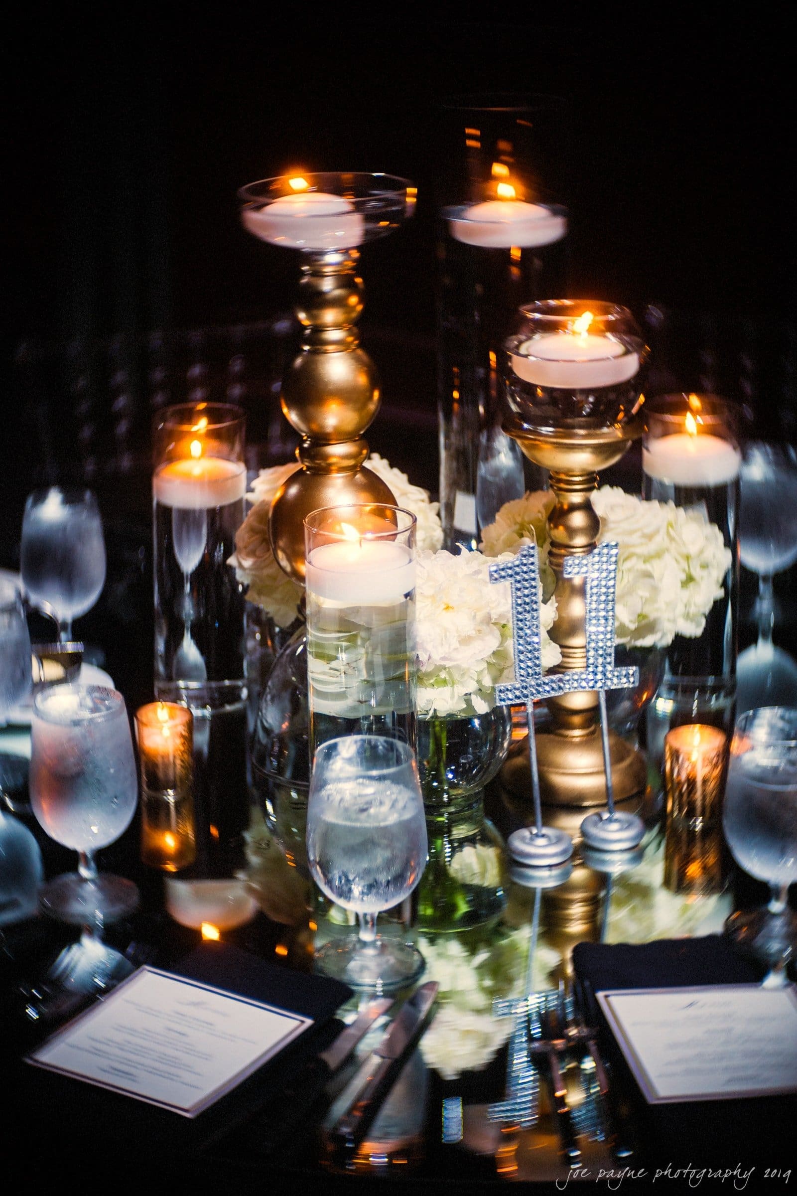 21c museum hotel durham wedding - kortne & terry A dimly lit elegant table setting with tall golden candle holders, white flowers, and several glasses of water. Plates on the table suggest a formal dining arrangement.