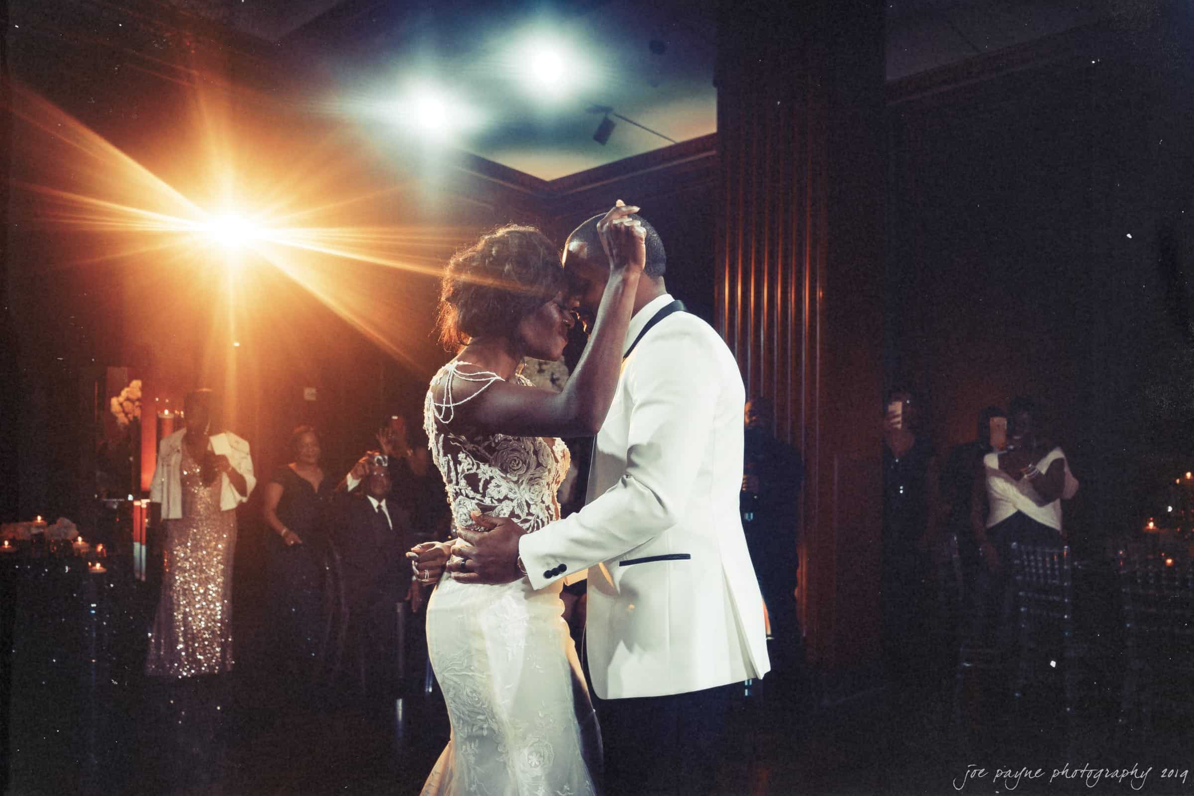 21c museum hotel durham wedding - kortne & terry A couple dances closely in an elegant setting, surrounded by dim lighting and guests. The woman wears a detailed lace gown, and the man is in a white tuxedo jacket. A bright light creates a soft, romantic atmosphere.