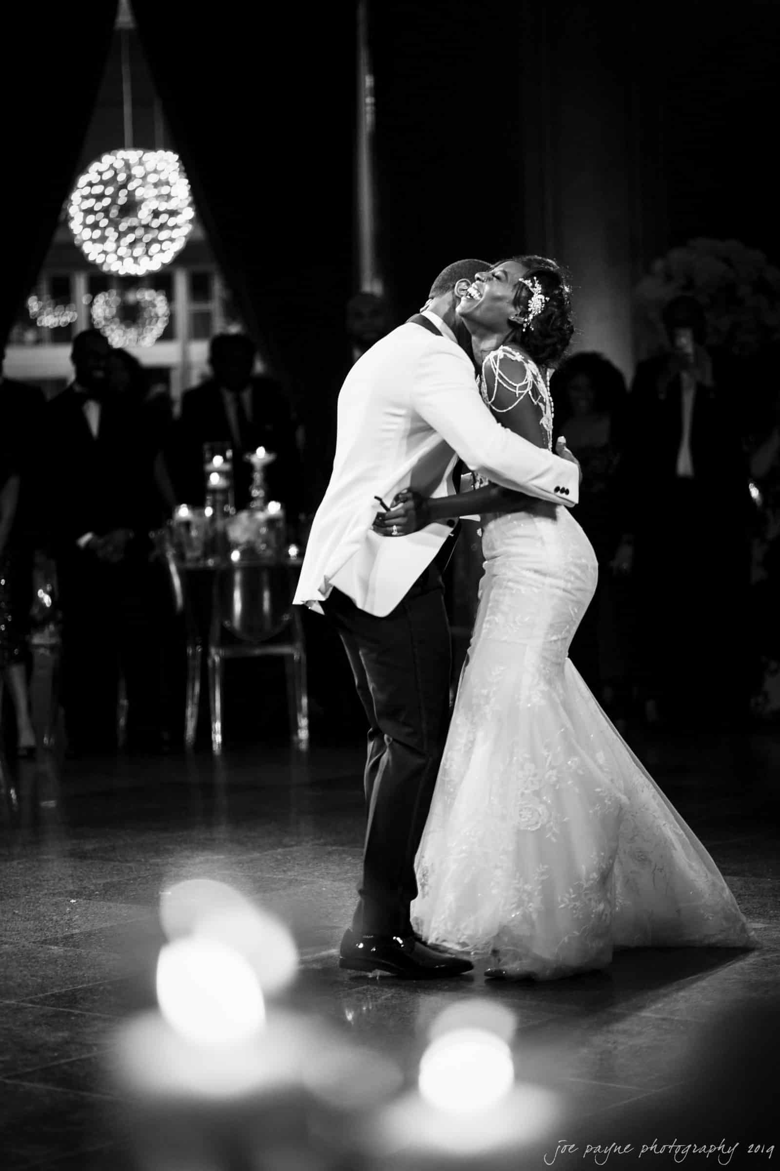 21c museum hotel durham wedding - kortne & terry A bride and groom share a romantic kiss on the dance floor, surrounded by wedding guests. The groom wears a white jacket, and the bride wears an elegant lace gown. The scene is captured in black and white, with soft lighting enhancing the moment.