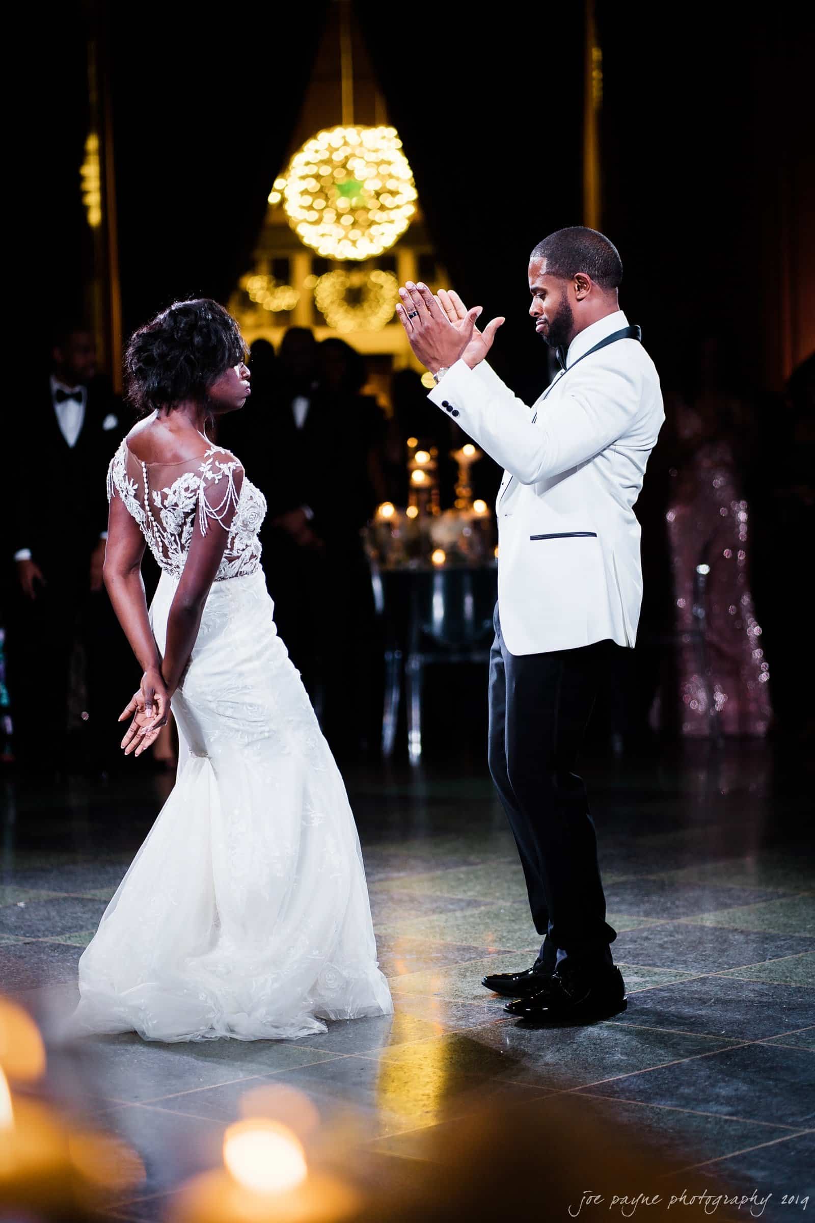 21c museum hotel durham wedding - kortne & terry A couple in formal attire dance together in a dimly lit ballroom. The woman wears a white gown with lace detailing, while the man wears a white tuxedo jacket and black pants. Warm lighting and a decorative chandelier create a festive atmosphere.