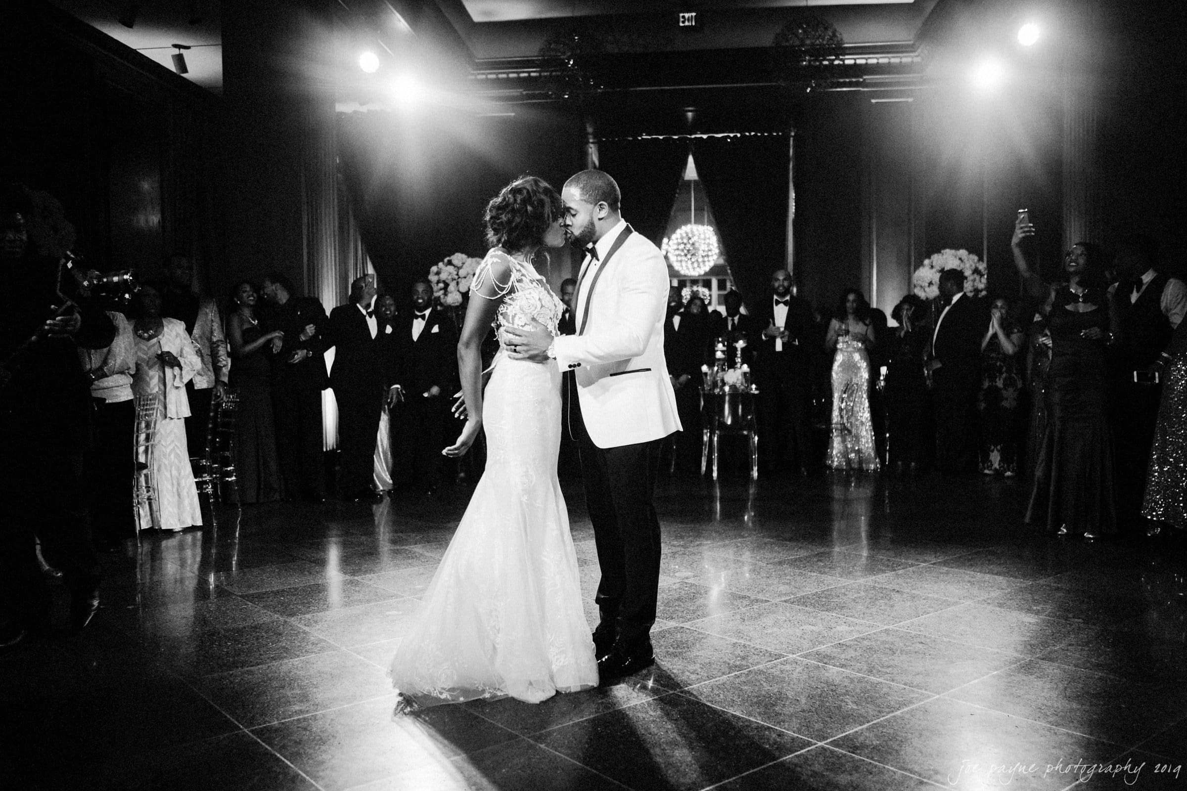 21c museum hotel durham wedding - kortne & terry A couple in formal attire dances closely on a polished floor in a dimly lit venue. Guests stand around them, watching the moment. The setting is elegant, with dramatic lighting and a chandelier in the background.