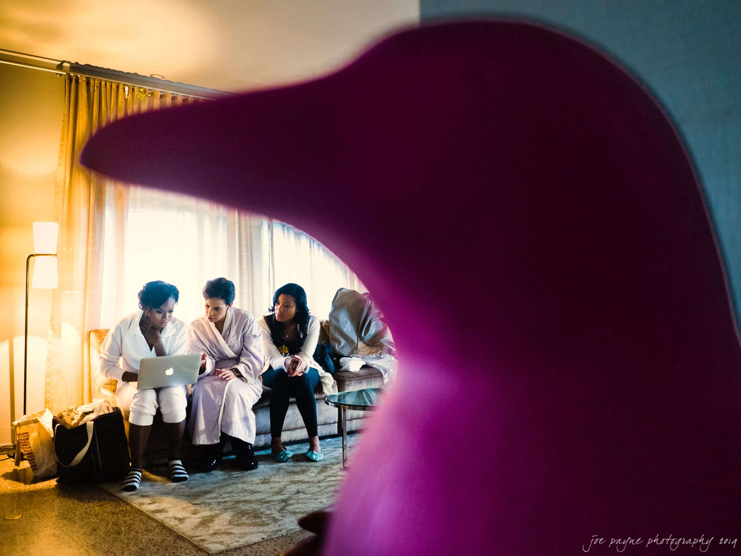 21c museum hotel durham wedding - kortne & terry A group of people sits on a couch, focused on a laptop. They are partially obscured by a large pink bird-shaped object in the foreground. The room has soft lighting from a standing lamp by the window.