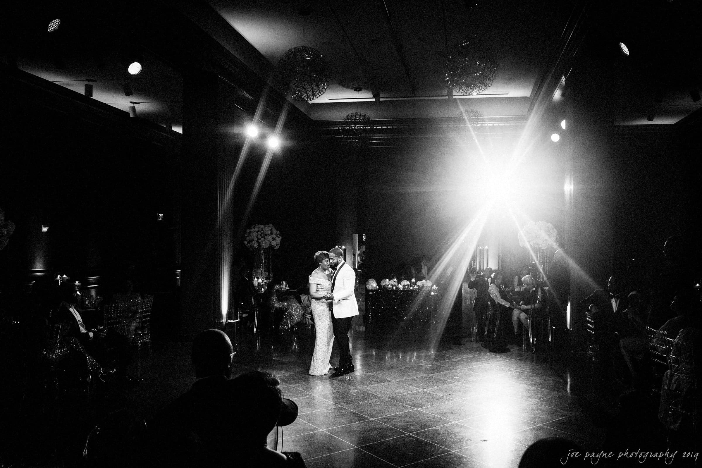 21c museum hotel durham wedding - kortne & terry A black and white photo of a couple dancing in a dimly lit ballroom. They are surrounded by guests seated at tables, with a bright light creating a dramatic effect. The scene exudes a romantic and elegant ambiance.