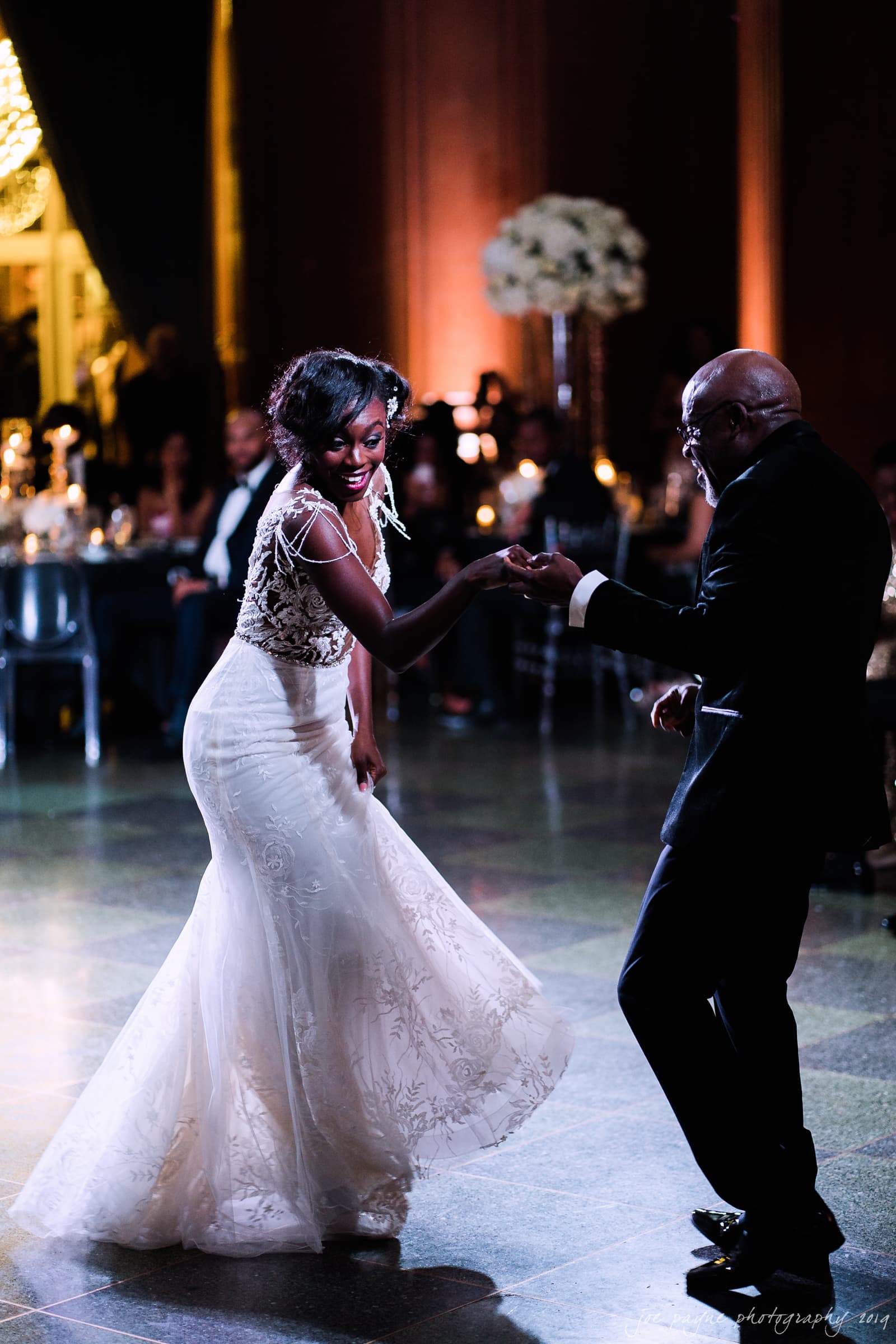 21c museum hotel durham wedding - kortne & terry A bride in a white dress joyfully dances with an older gentleman in a suit at a nighttime event. The room is warmly lit with candles and guests seated in the background, creating an elegant and festive atmosphere.