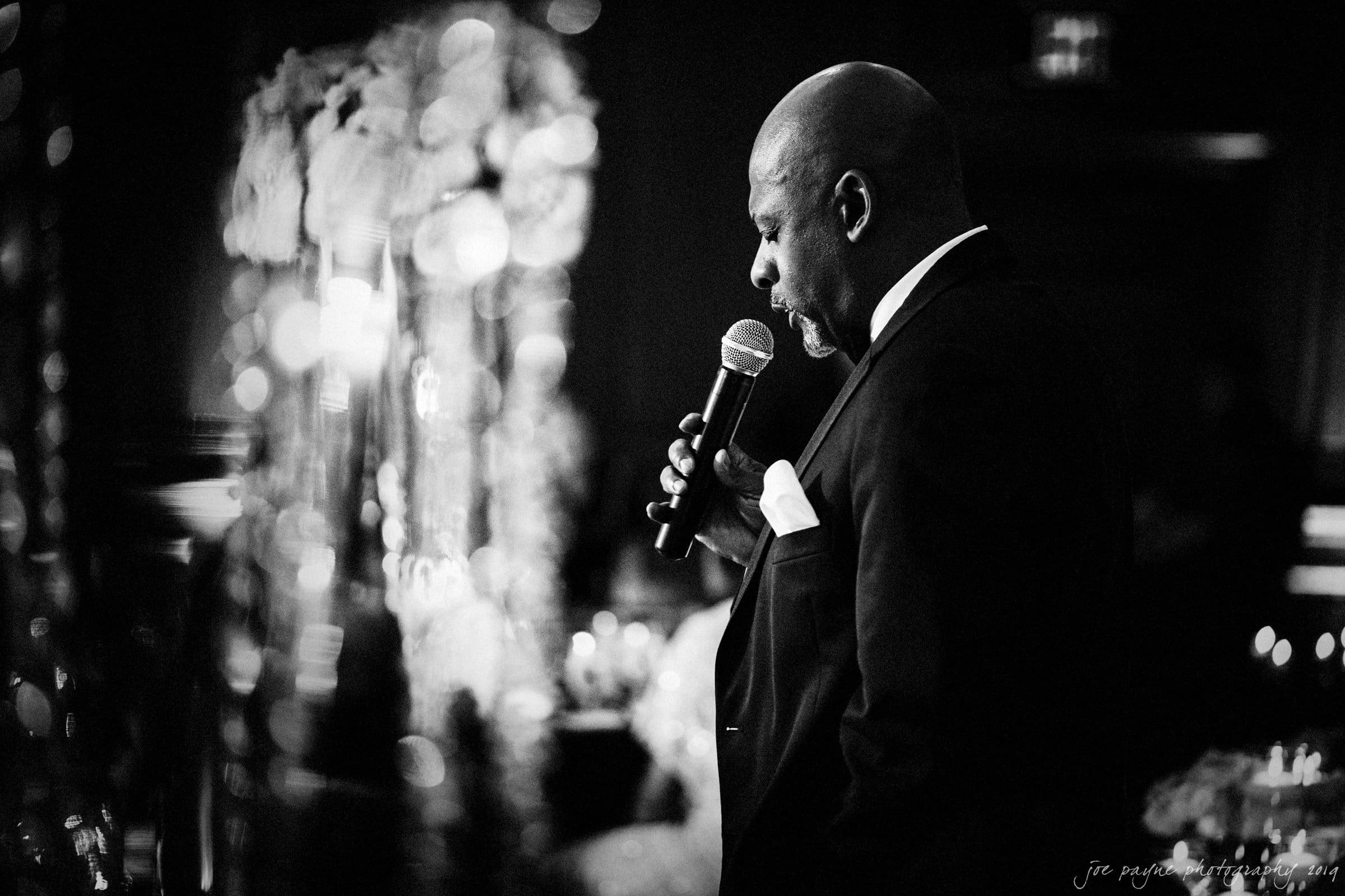 21c museum hotel durham wedding - kortne & terry A man in a suit speaks into a microphone at an event. He is captured in profile, and the background is softly blurred with decorative elements. The scene has a black-and-white, intimate atmosphere.