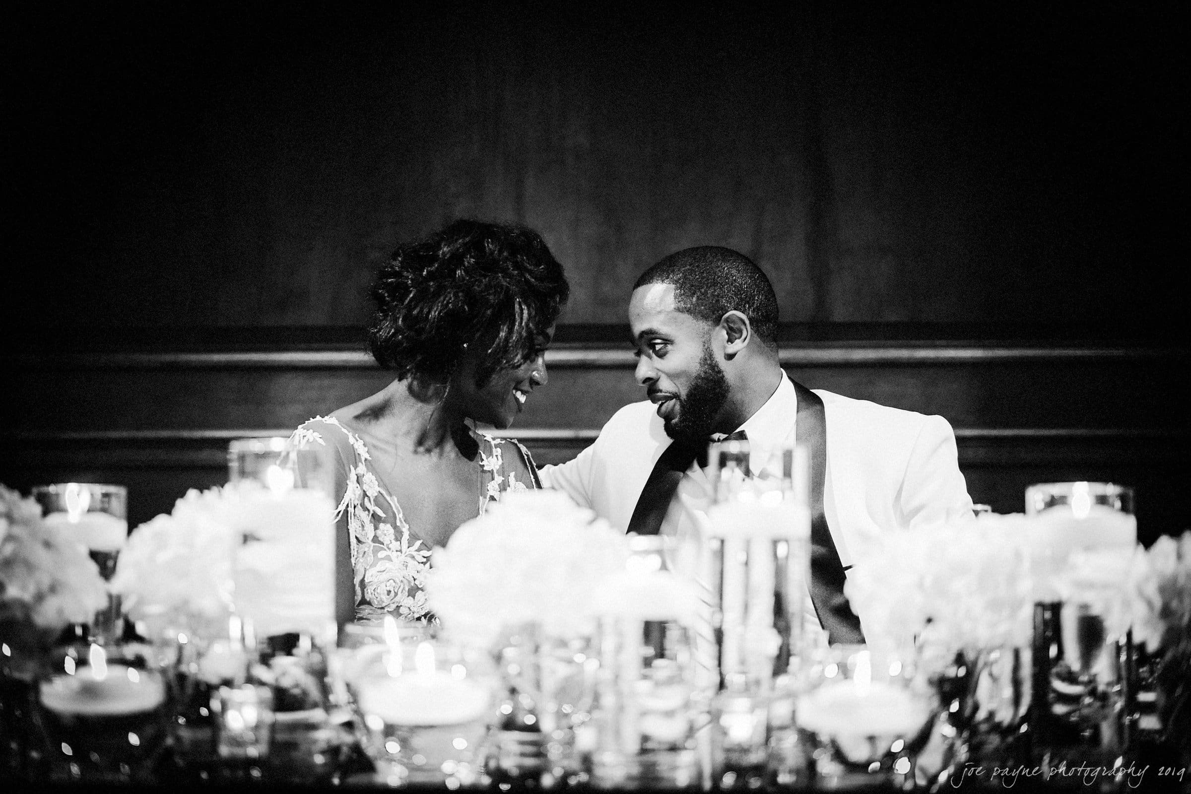 21c museum hotel durham wedding - kortne & terry A black and white photo of a couple sitting closely at an elegantly decorated table adorned with flowers and candles. They are leaning toward each other, sharing a warm, intimate moment.
