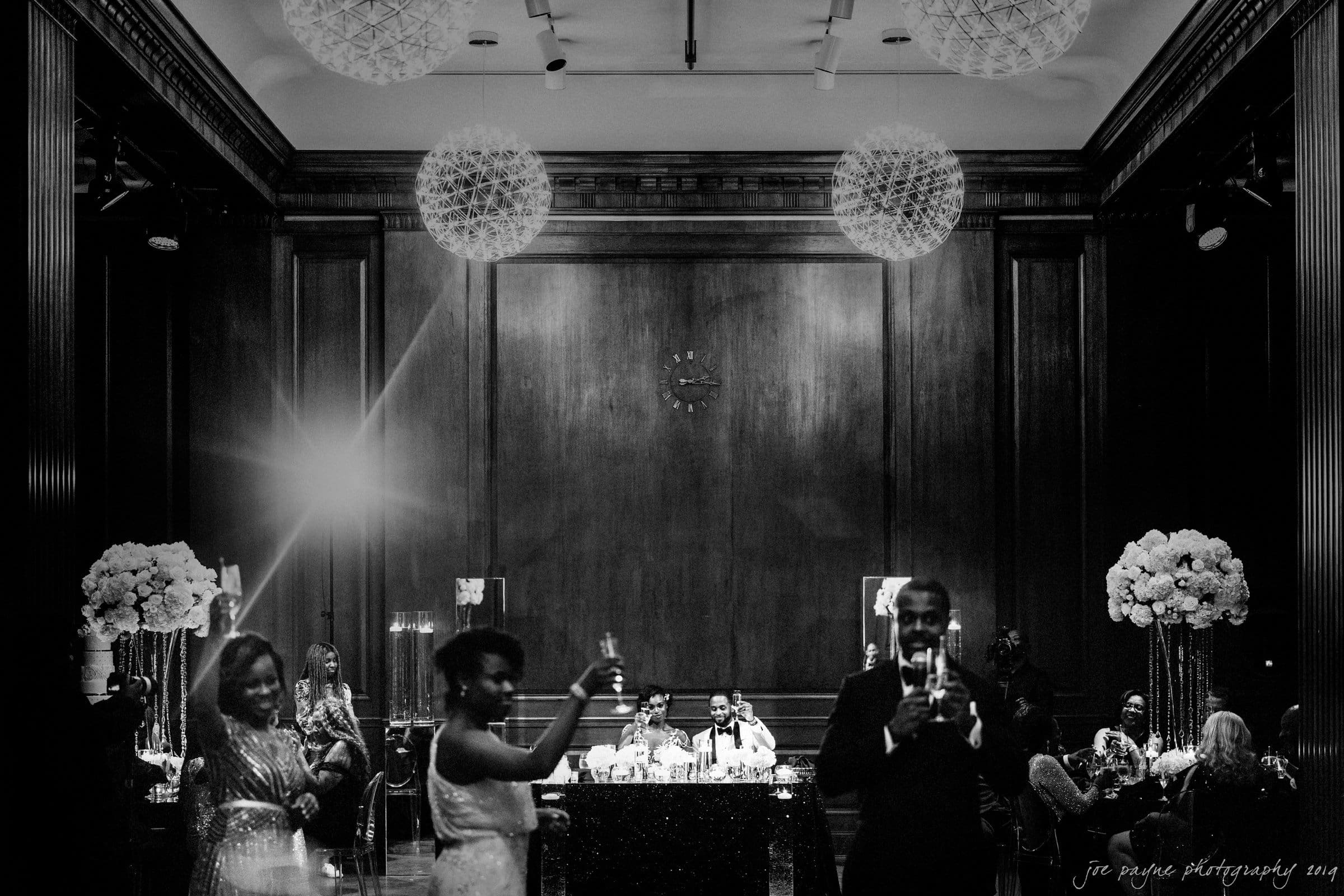 21c museum hotel durham wedding - kortne & terry A black and white image of a formal event in an elegant room. Several people raise glasses in a toast. Three large chandeliers hang from the ceiling, and a panelled wall serves as the backdrop. A couple sits at a table in the center background.