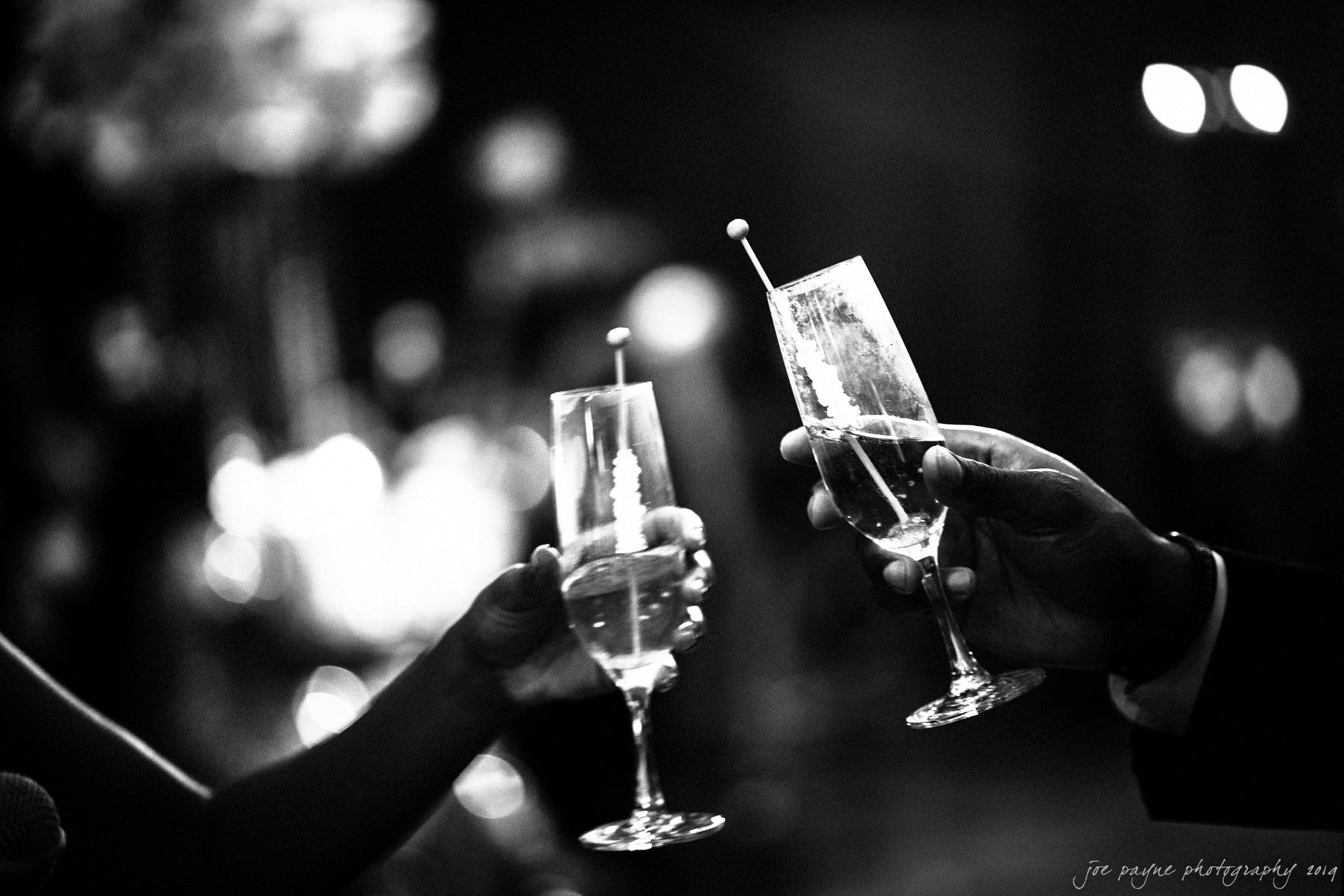 21c museum hotel durham wedding - kortne & terry Black and white photo of two people clinking champagne flutes in a celebratory toast, with a soft-focus background and subtle bokeh lights.