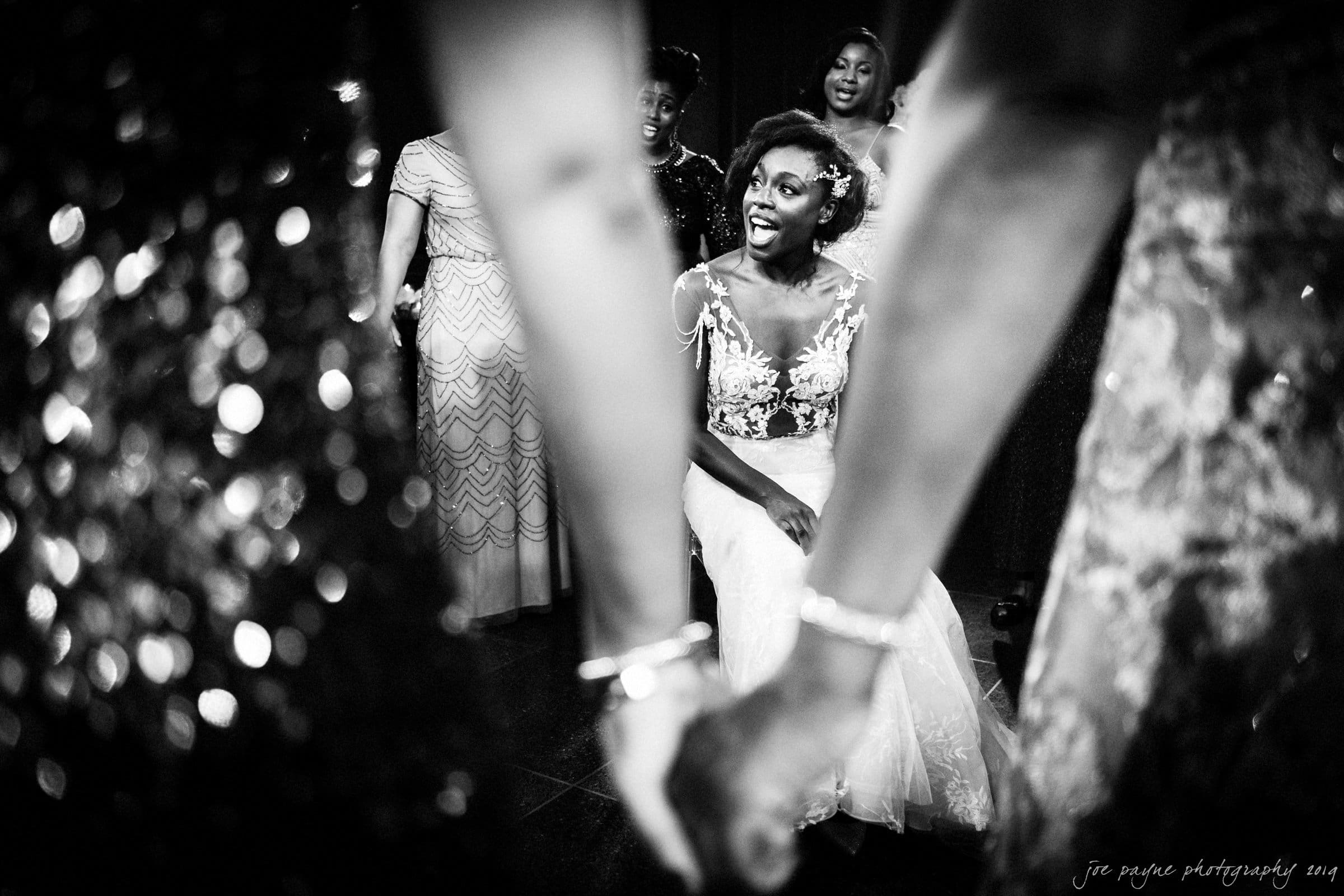 21c museum hotel durham wedding - kortne & terry A joyful bride sits and laughs, surrounded by bridesmaids. The scene is captured in black and white, highlighting their sparkling dresses. Two women hold hands in the foreground, framing the bride with their arms.
