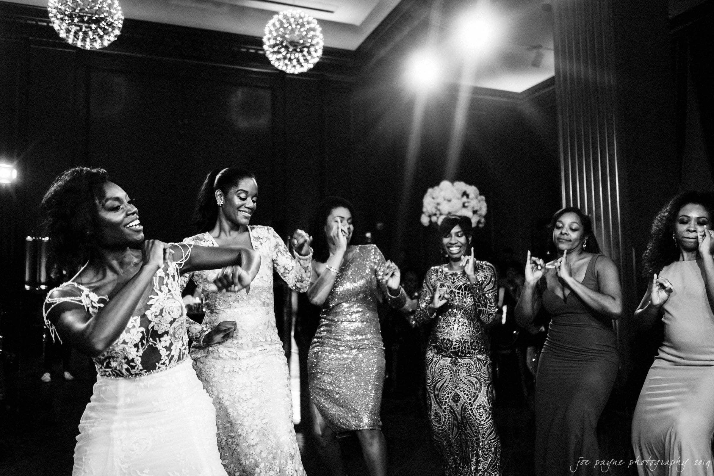 21c museum hotel durham wedding - kortne & terry A group of women in elegant dresses joyfully dancing in a dimly lit room. The atmosphere is lively, and they are smiling and engaged in the moment. The ceiling is adorned with spherical lights adding to the festive ambiance.