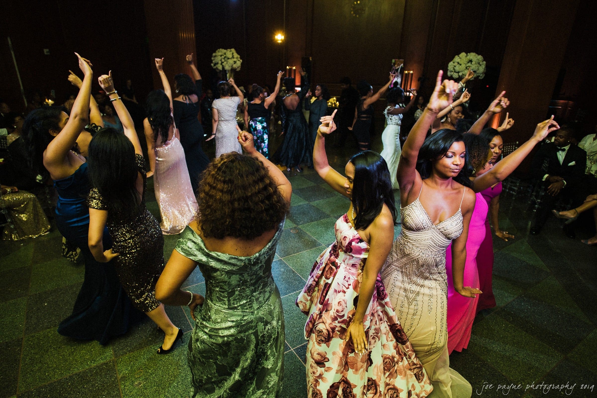 21c museum hotel durham wedding - kortne & terry A group of people dance in a circle on a dimly lit dance floor, wearing elegant dresses in various colors. The atmosphere is lively and celebratory, with floral decorations in the background.
