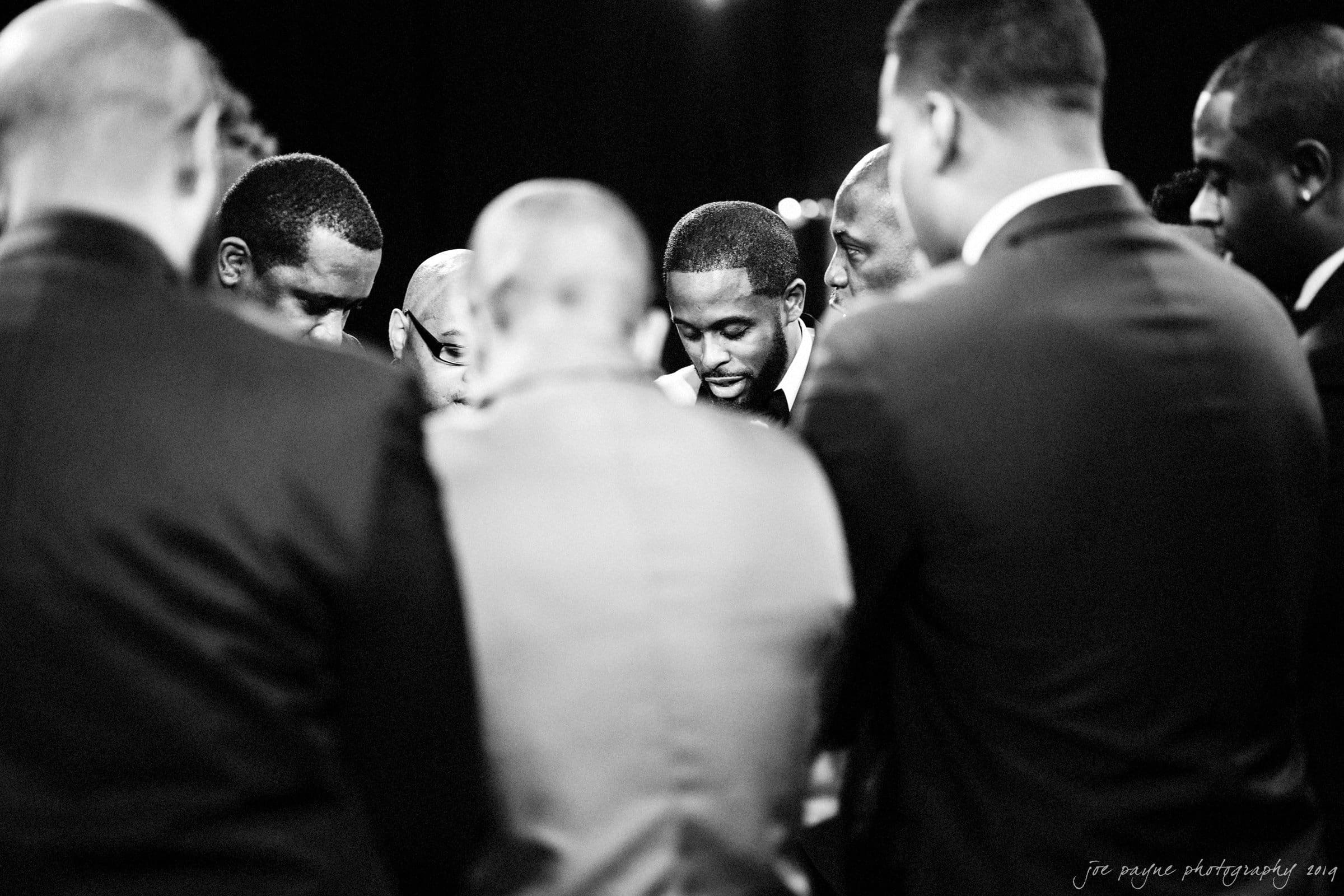 21c museum hotel durham wedding - kortne & terry A group of men in suits stand closely together in a circle, appearing to be engaged in a moment of reflection or prayer. The image is in black and white, with expressions of focus and solemnity visible on their faces.
