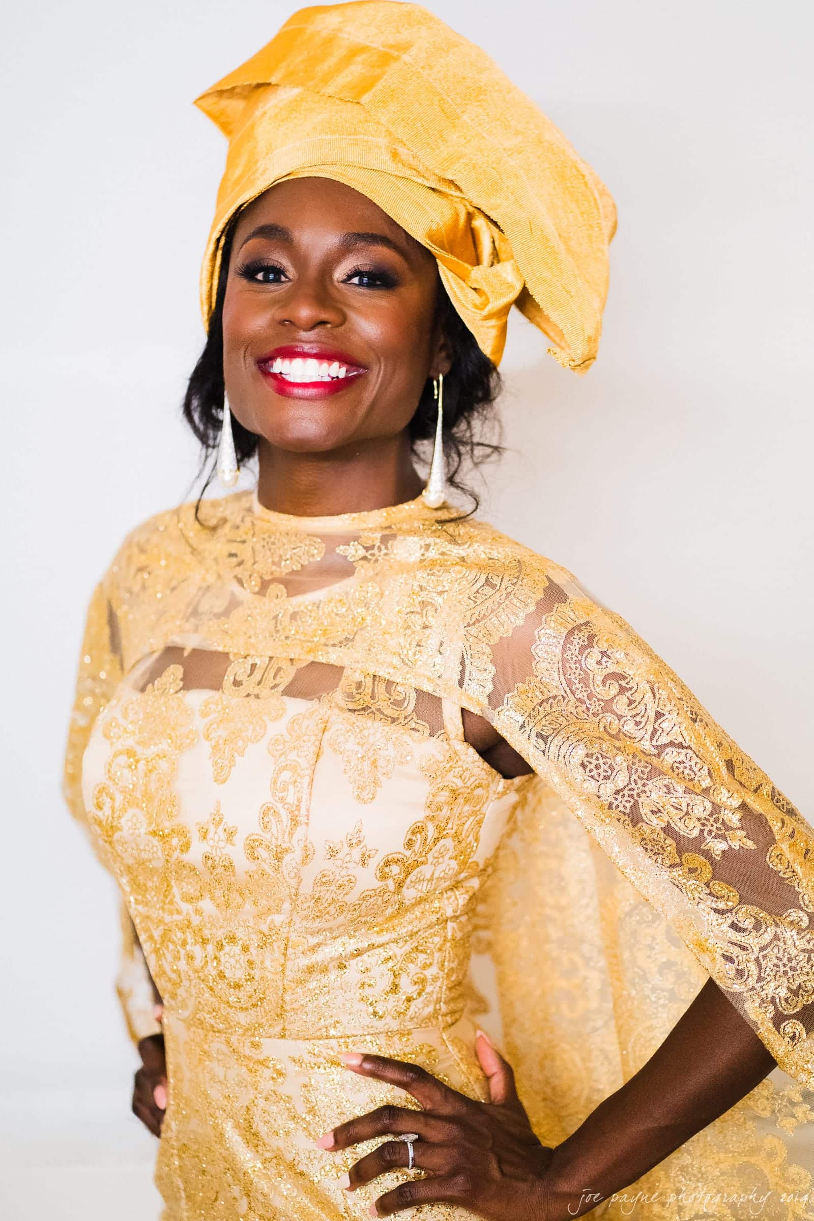 21c museum hotel durham wedding - kortne & terry A woman smiles confidently wearing a vibrant gold lace dress with a matching headwrap. She stands with her hands on her hips, exuding elegance and grace.