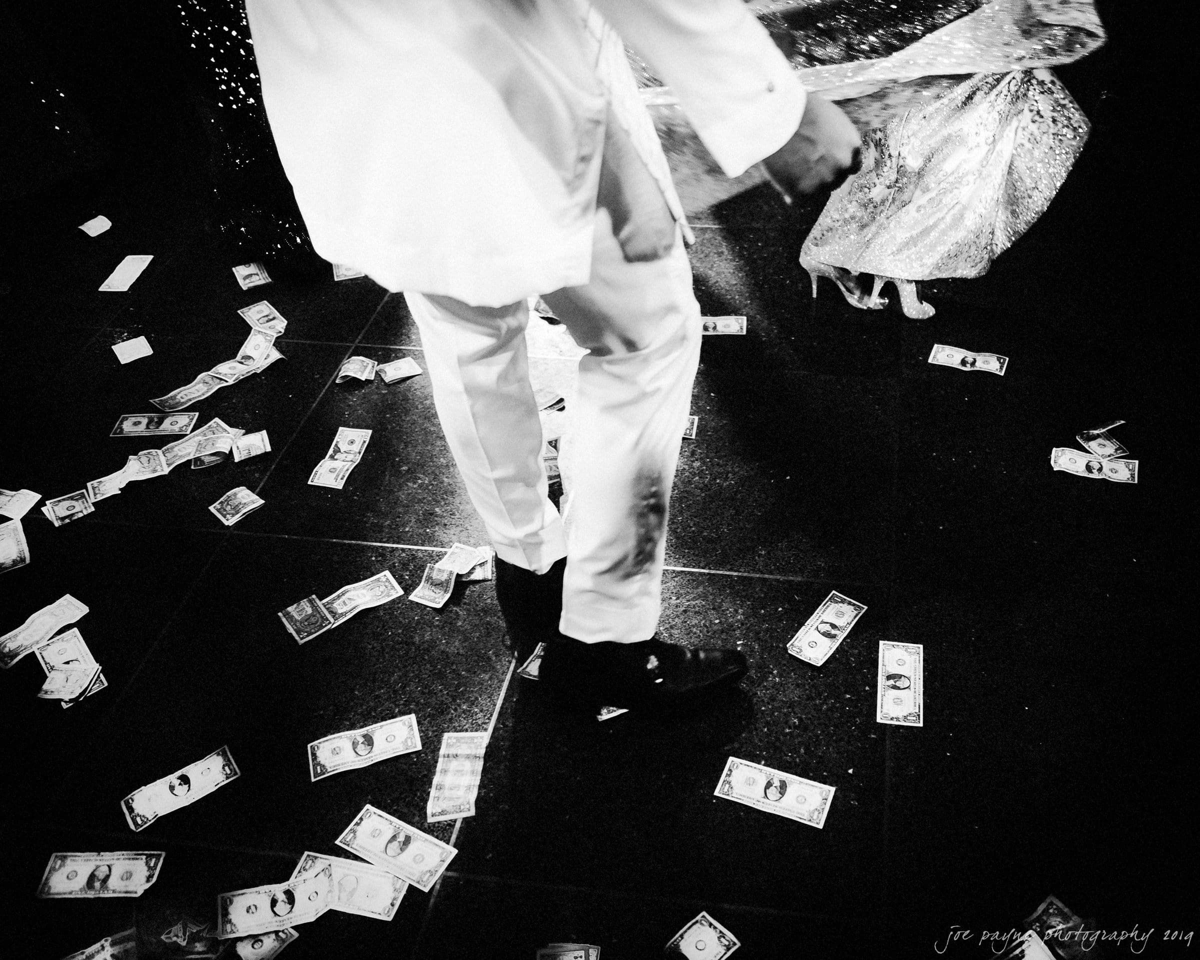 21c museum hotel durham wedding - kortne & terry A black-and-white photo showing a person in a white suit dancing on a floor covered with scattered paper money. Another person in a sparkly outfit is partially visible, creating a dynamic, lively atmosphere.