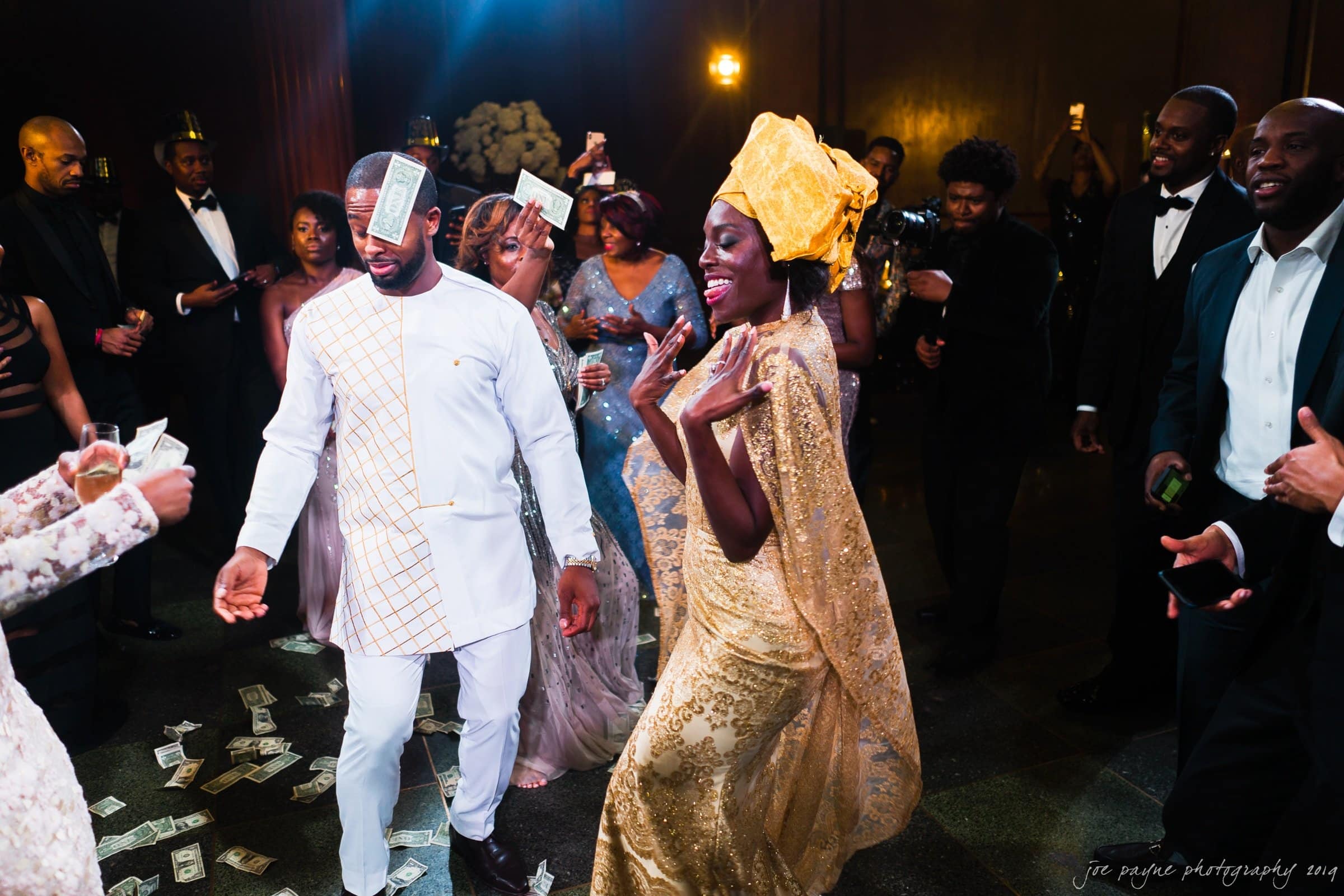 21c museum hotel durham wedding - kortne & terry A joyful couple dances energetically at a celebration. The man wears an embroidered white outfit with money stuck to his forehead, and the woman is in a golden dress and headwrap. People surround them, tossing money and enjoying the festive atmosphere.