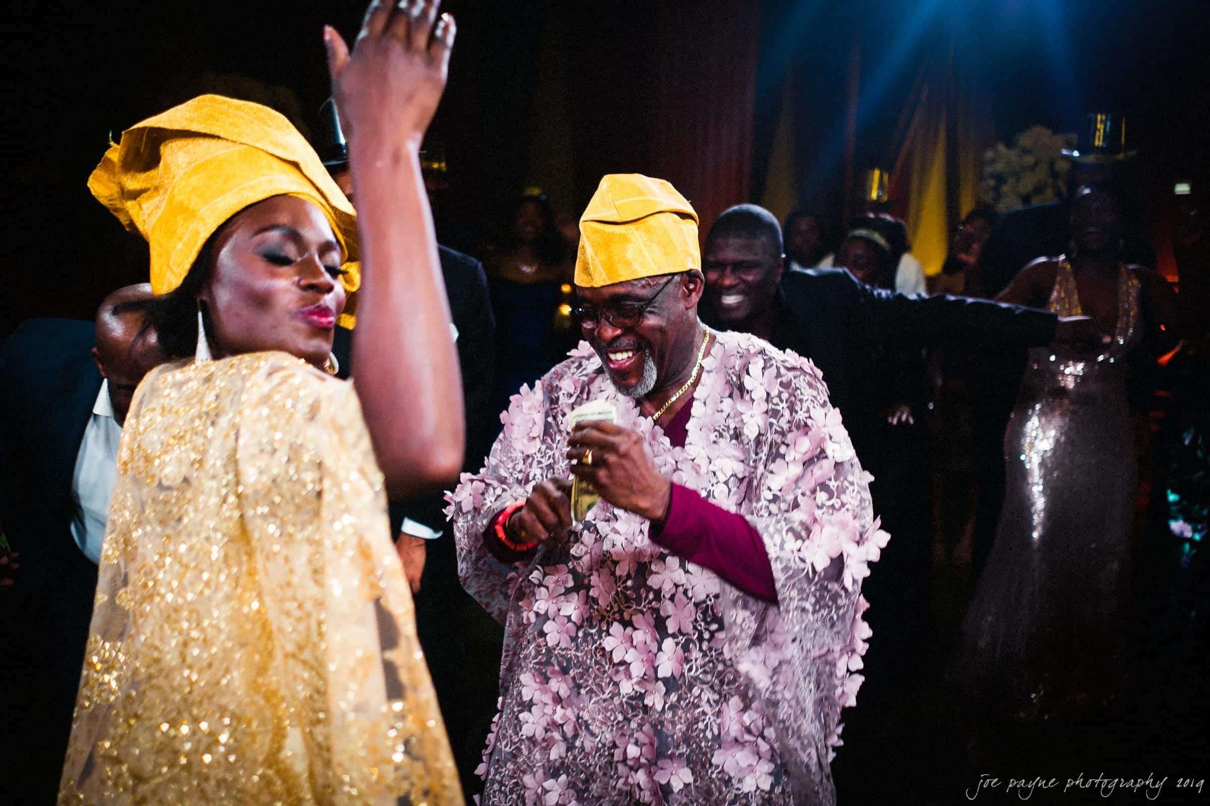 21c museum hotel durham wedding - kortne & terry A lively celebration with people dancing joyfully. A woman and a man in vibrant traditional attire, including yellow headwraps and a floral-patterned cloak, are at the center, smiling and holding drinks. The background features more people and festive lighting.