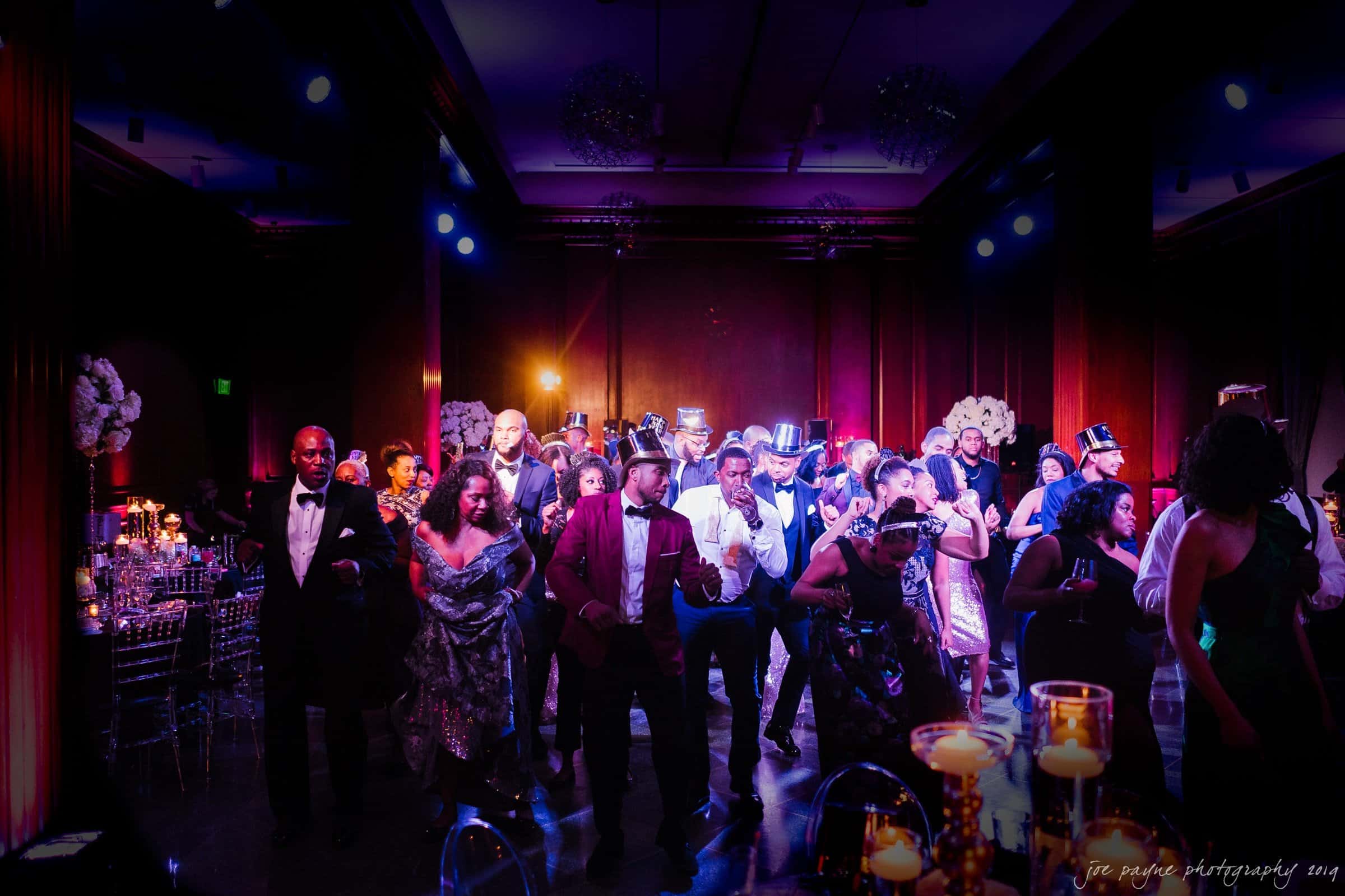 21c museum hotel durham wedding - kortne & terry A lively group of people dressed in formal attire dance on a dimly lit ballroom floor. The room is decorated with white floral arrangements and glowing candles, while colored stage lights illuminate the scene.
