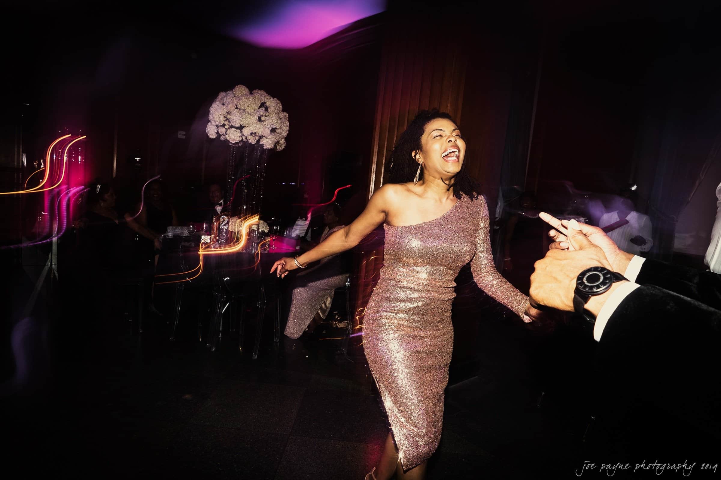 21c museum hotel durham wedding - kortne & terry A woman in a sparkling one-shoulder dress joyfully dances at a vibrant event. She is surrounded by blurred lights and seated guests. Someones hand is visible pointing in her direction.