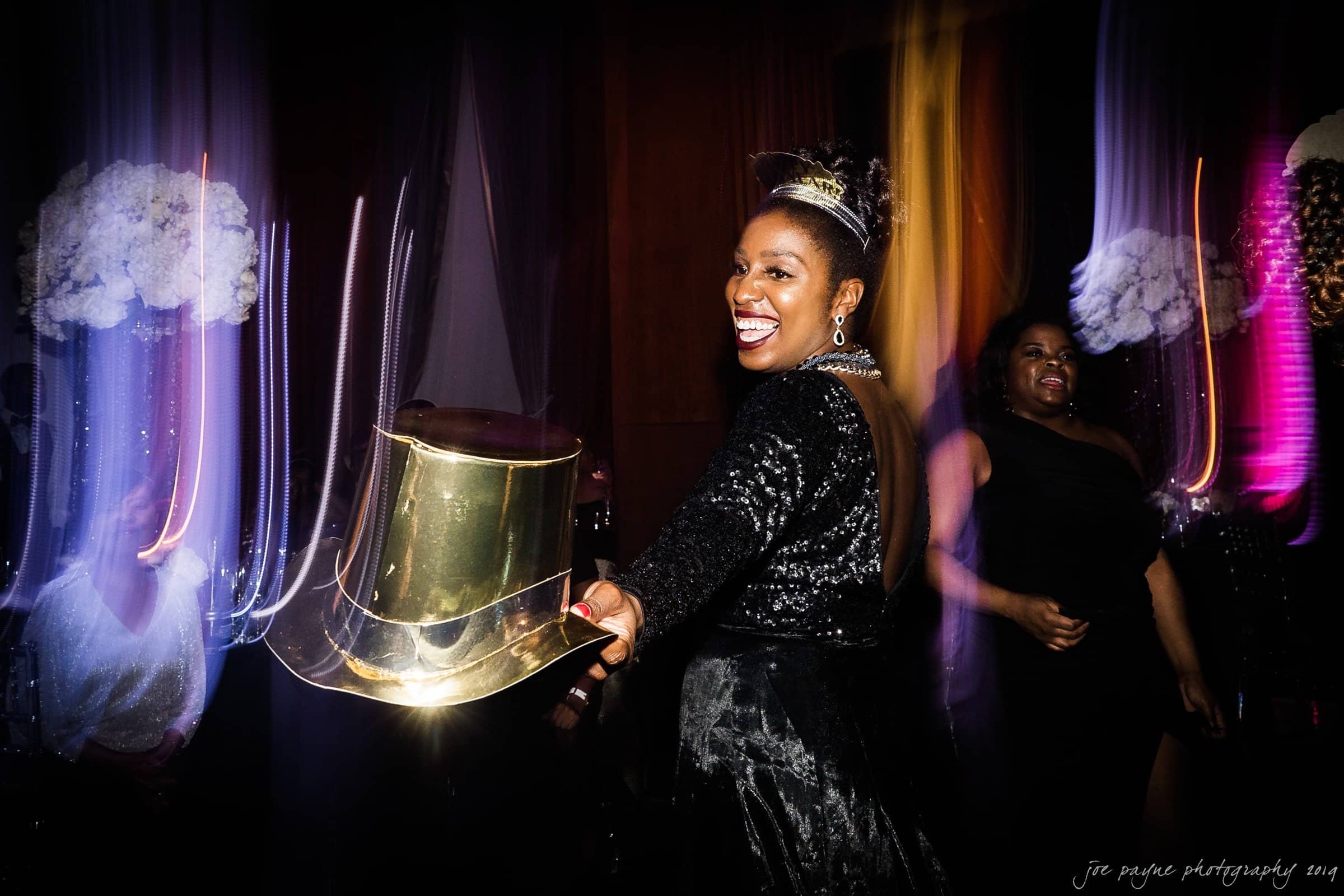 21c museum hotel durham wedding - kortne & terry A woman in a sparkling black outfit and tiara smiles brightly, holding a gold top hat. She stands in a dimly lit room with blurred colorful lights and floral decorations in the background.