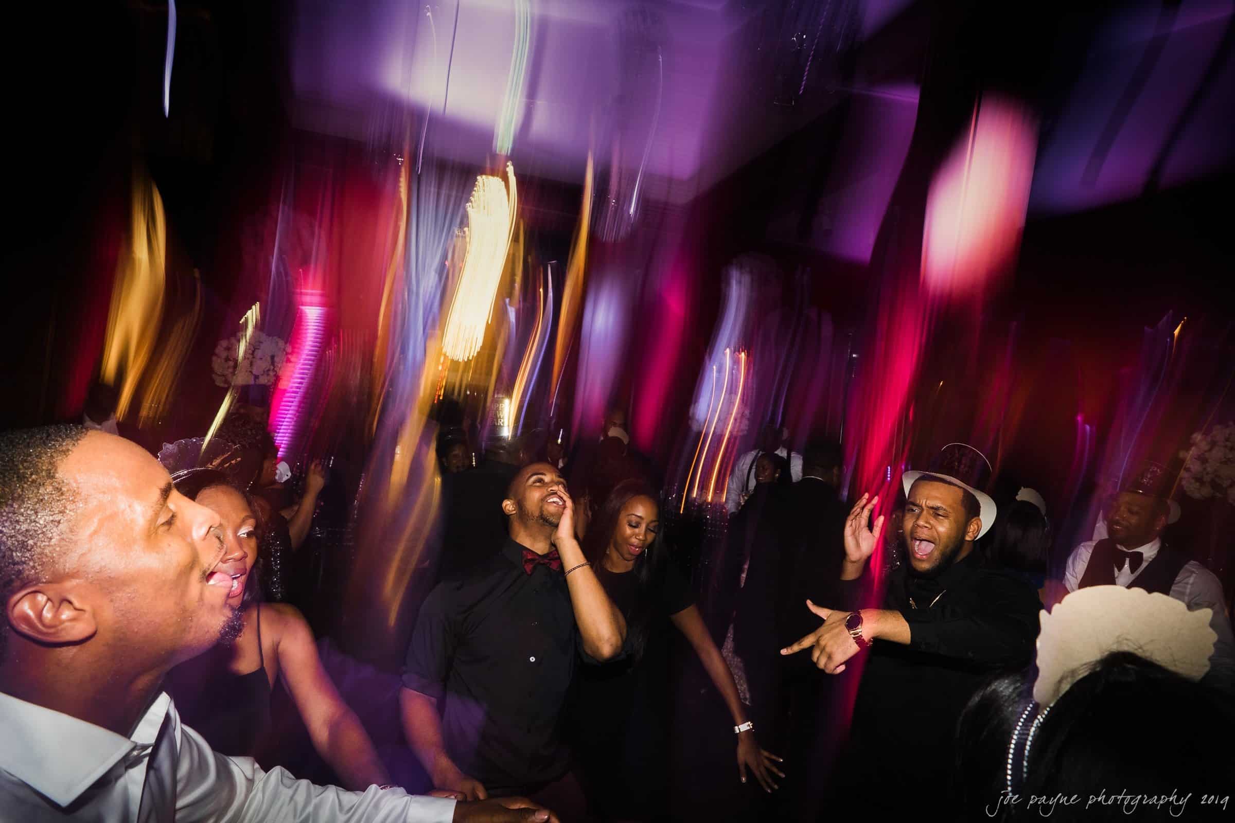 21c museum hotel durham wedding - kortne & terry A lively group of people dance energetically under vibrant, colorful lights. Motion blur adds a dynamic feel to the scene, with individuals clearly enjoying the music and moment in a festive atmosphere.