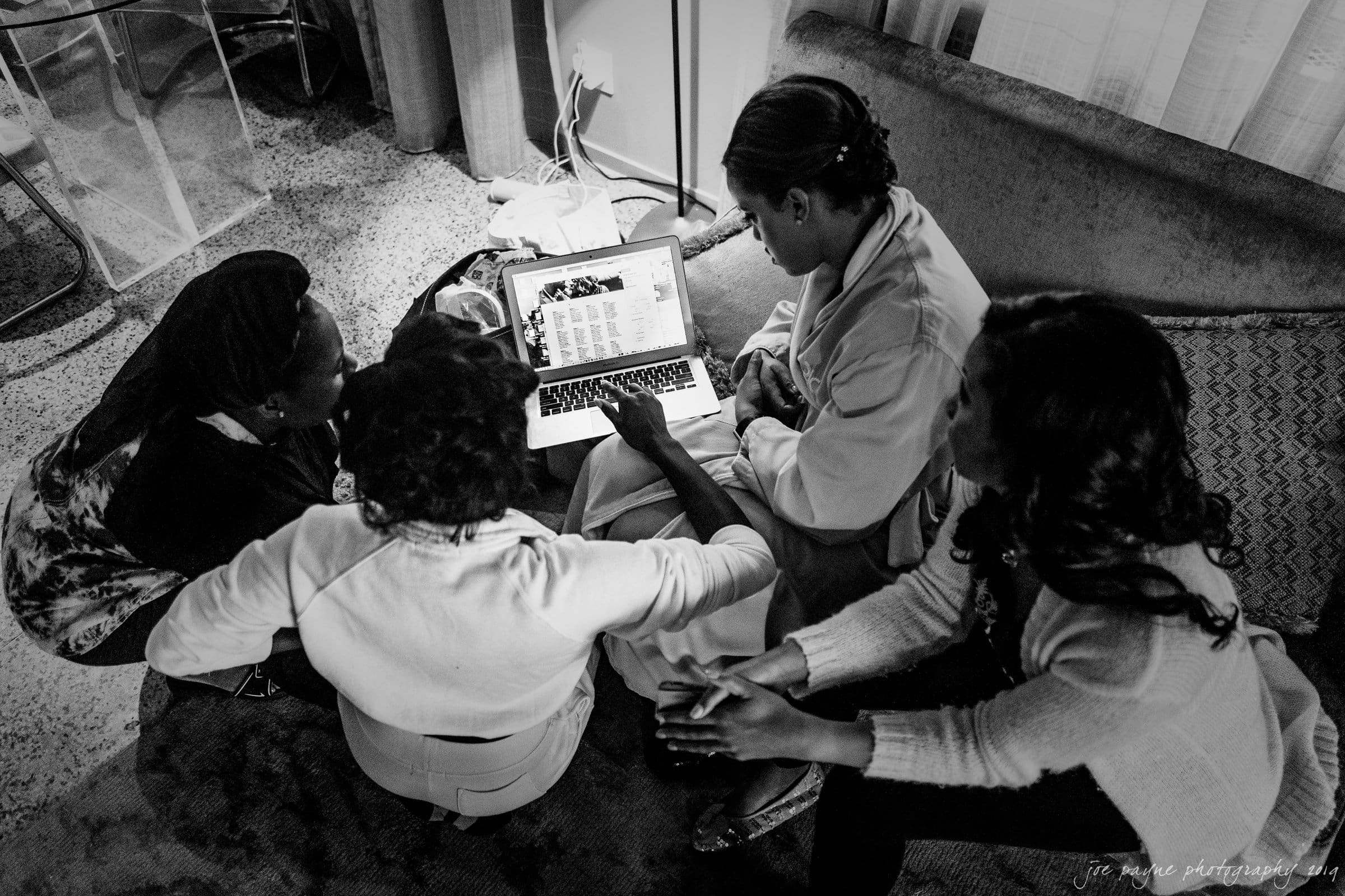 21c museum hotel durham wedding - kortne & terry A black and white image of four women sitting on a couch, gathered around a laptop. One woman points at the screen while the others watch attentively. The room has a cozy atmosphere with dim lighting.