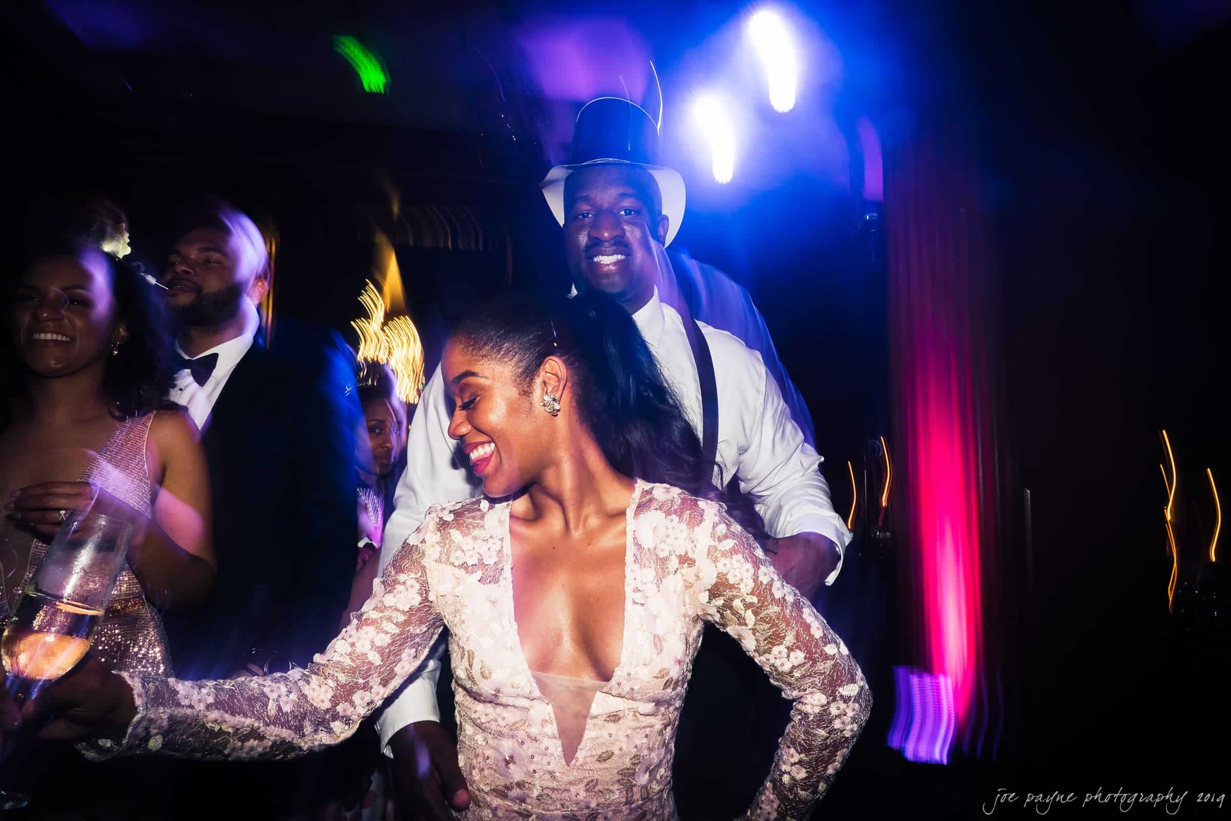 21c museum hotel durham wedding - kortne & terry A lively party scene with elegantly dressed guests dancing. A woman in a lacy dress holds a drink and smiles, while a man in a top hat dances behind her. Colorful lights create a vibrant atmosphere.