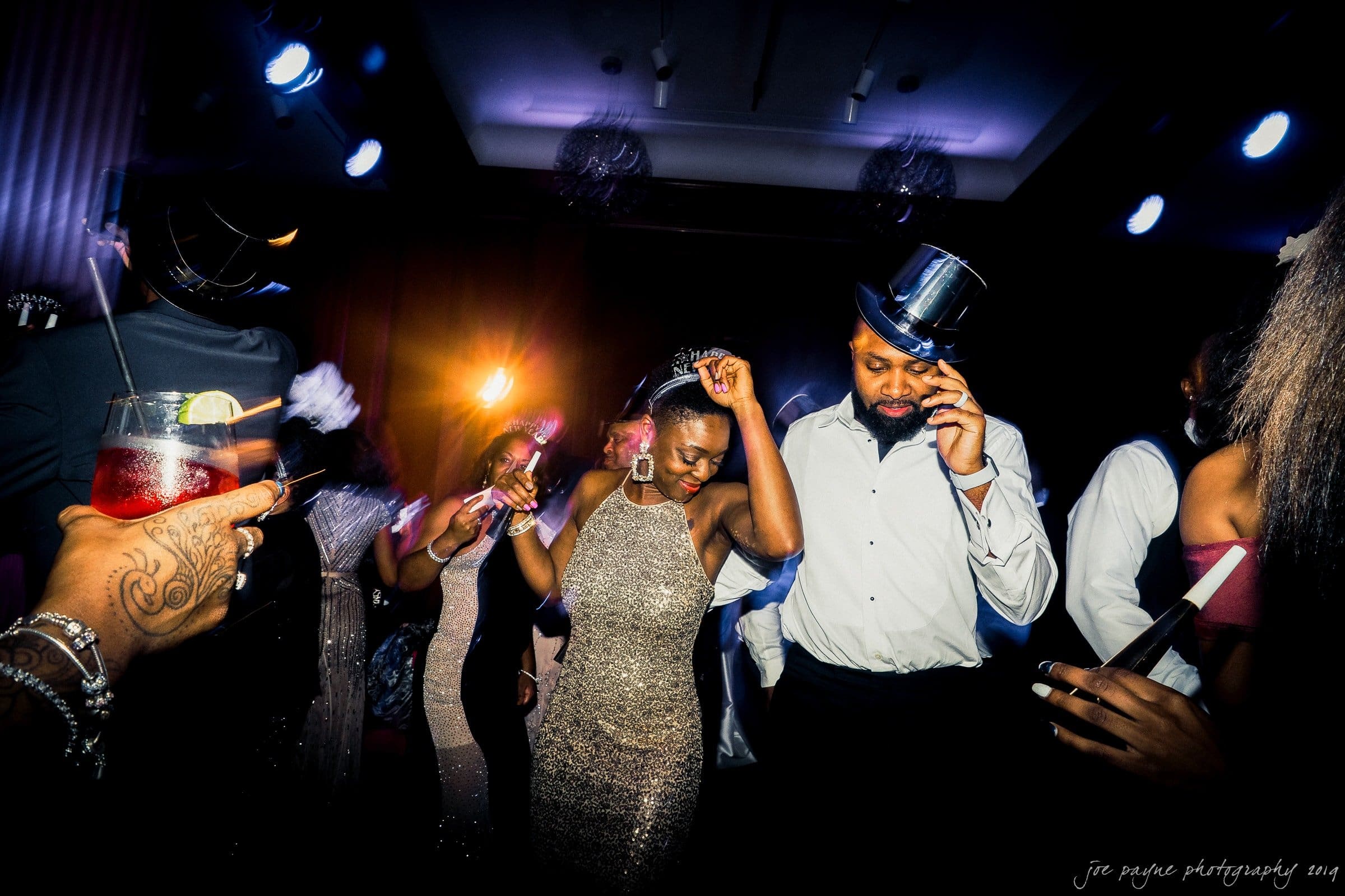 21c museum hotel durham wedding - kortne & terry A group of people dance energetically under bright lights at a party. A woman in a glittery dress and a man wearing a top hat are prominently in the center. Others are enjoying drinks and the lively atmosphere.