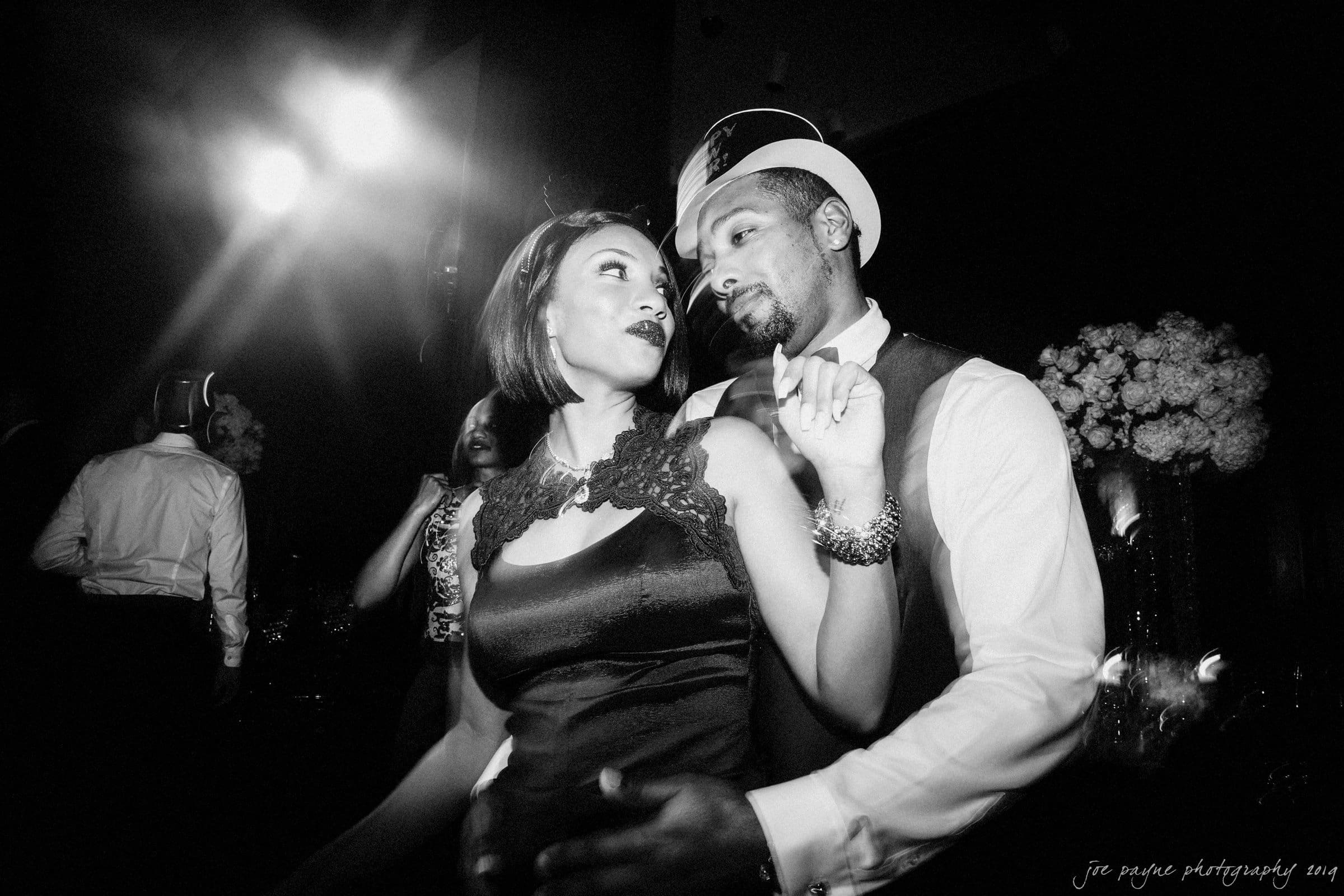 21c museum hotel durham wedding - kortne & terry Black and white photo of a couple dancing closely at a formal event. The woman is in a dark dress with lace detail, and the man is wearing a vest and hat. They seem to be gazing at each other amidst a lively atmosphere.