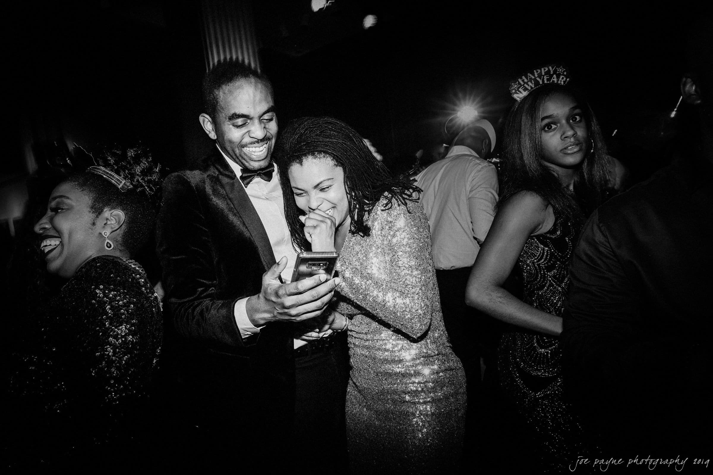 21c museum hotel durham wedding - kortne & terry A group of people in formal attire enjoy a New Years celebration. A man and woman in the center share a laugh while looking at a phone. A woman with a Happy New Year headband stands nearby. The setting is dark with festive lighting.