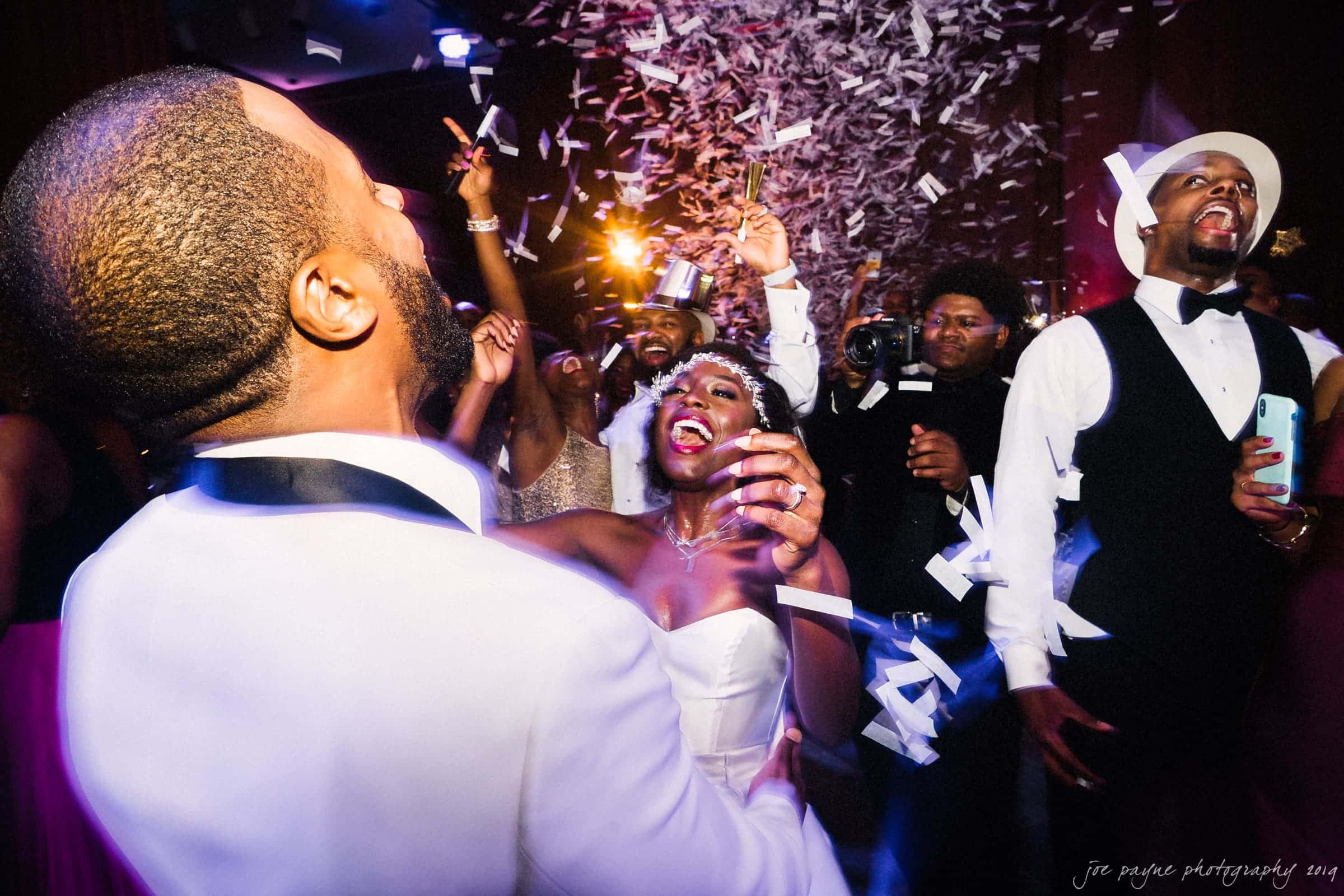 21c museum hotel durham wedding - kortne & terry A joyful wedding celebration with a bride and groom laughing as confetti rains down. Guests are cheering, clapping, and capturing the moment with phones and cameras. The vibrant scene conveys excitement and happiness.