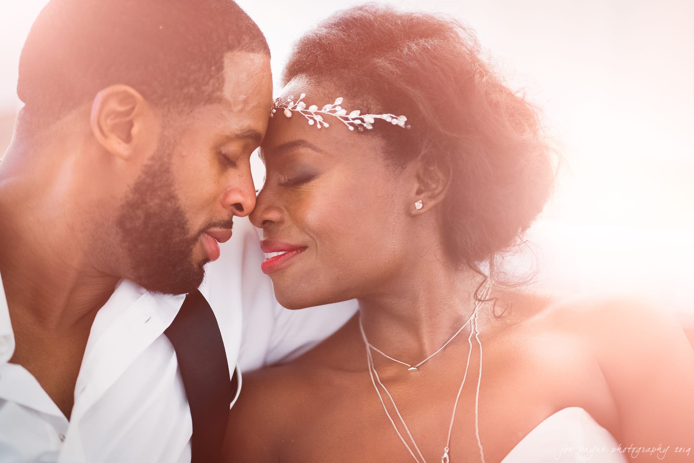 Close-up of a couple embracing tenderly, their foreheads touching. The woman wears a delicate headpiece and necklace, while the man is in a partially unbuttoned shirt. They both have serene expressions, with soft, warm lighting enveloping them.
