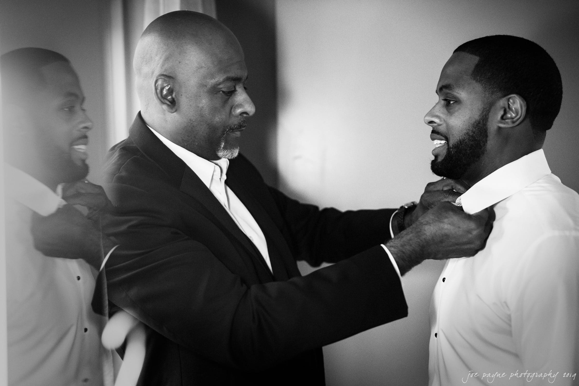 21c museum hotel durham wedding - kortne & terry A black-and-white image of an older man helping a younger man adjust his tie. Both are wearing suits, and the atmosphere is intimate and focused, suggesting a special occasion or event.