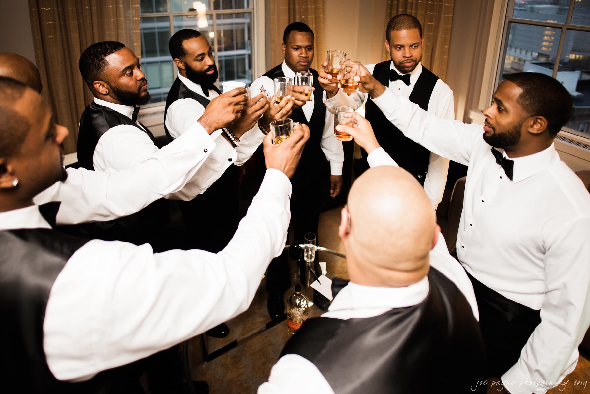 21c museum hotel durham wedding - kortne & terry A group of seven men dressed in formal attire, including white shirts and black vests, stand in a circle raising glasses in a toast. They appear to be in a well-lit room with large windows, celebrating an occasion.