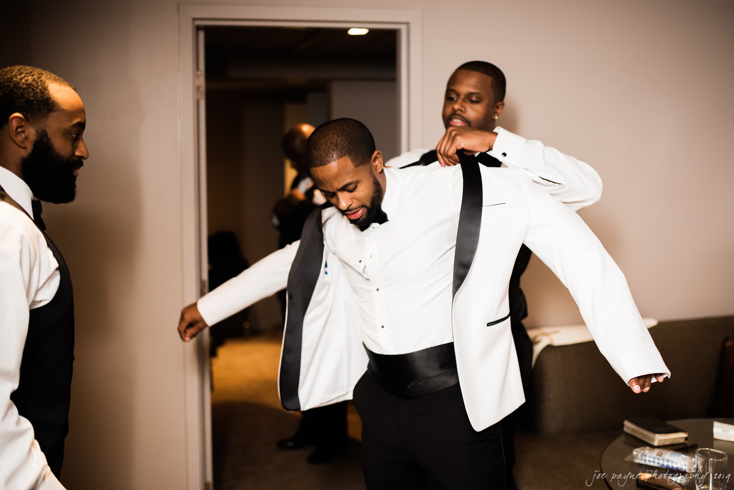21c museum hotel durham wedding - kortne & terry A man assists another man in putting on a white tuxedo jacket. Both are dressed formally, with bow ties, in a room with soft lighting. Another man in the foreground observes, and a fourth person is partially visible in the background.