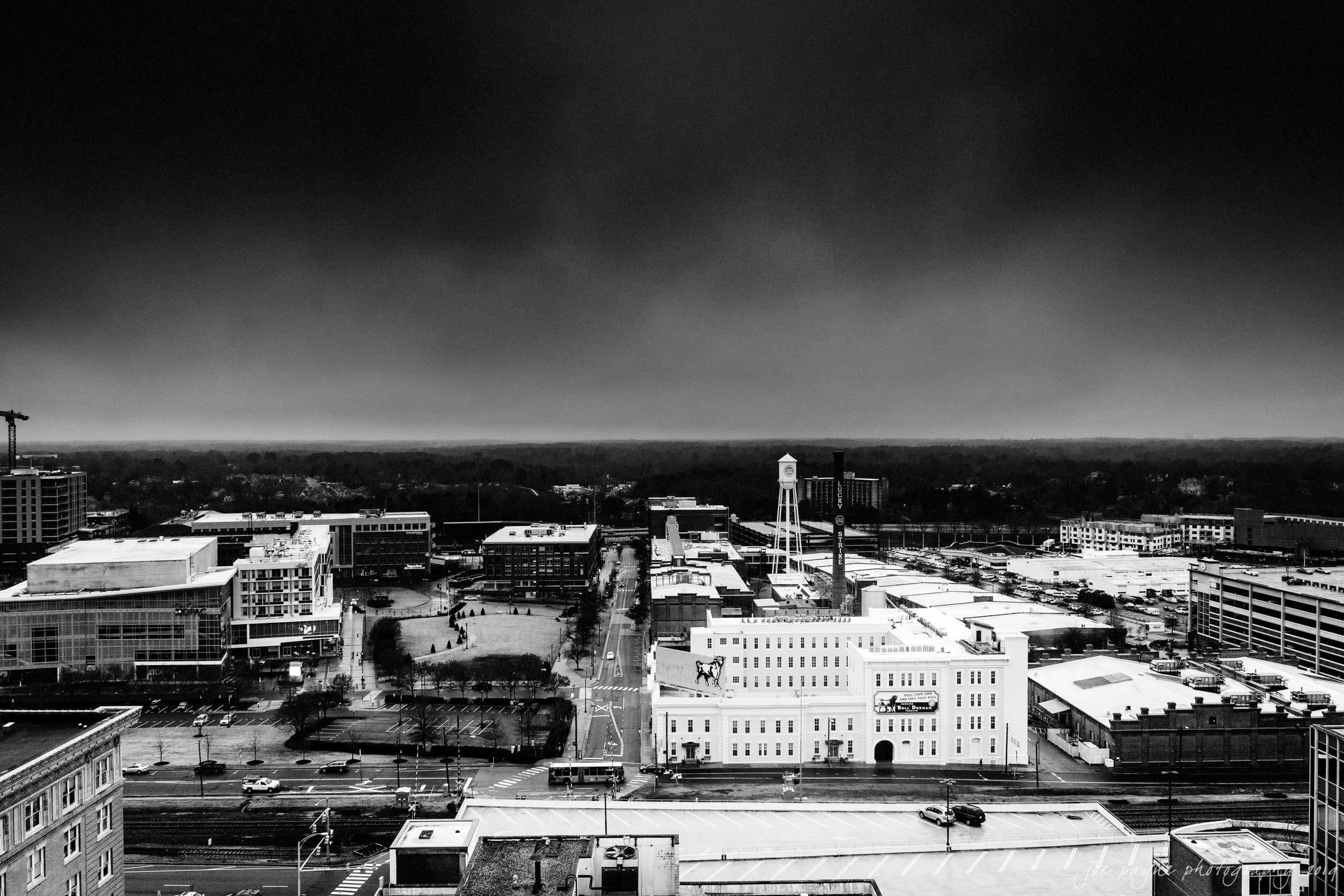 21c museum hotel durham wedding - kortne & terry A dramatic black and white aerial view of a cityscape under cloudy skies. Prominent buildings include a tall white structure and a water tower. Streets and parking areas are visible, contributing to a moody urban atmosphere.