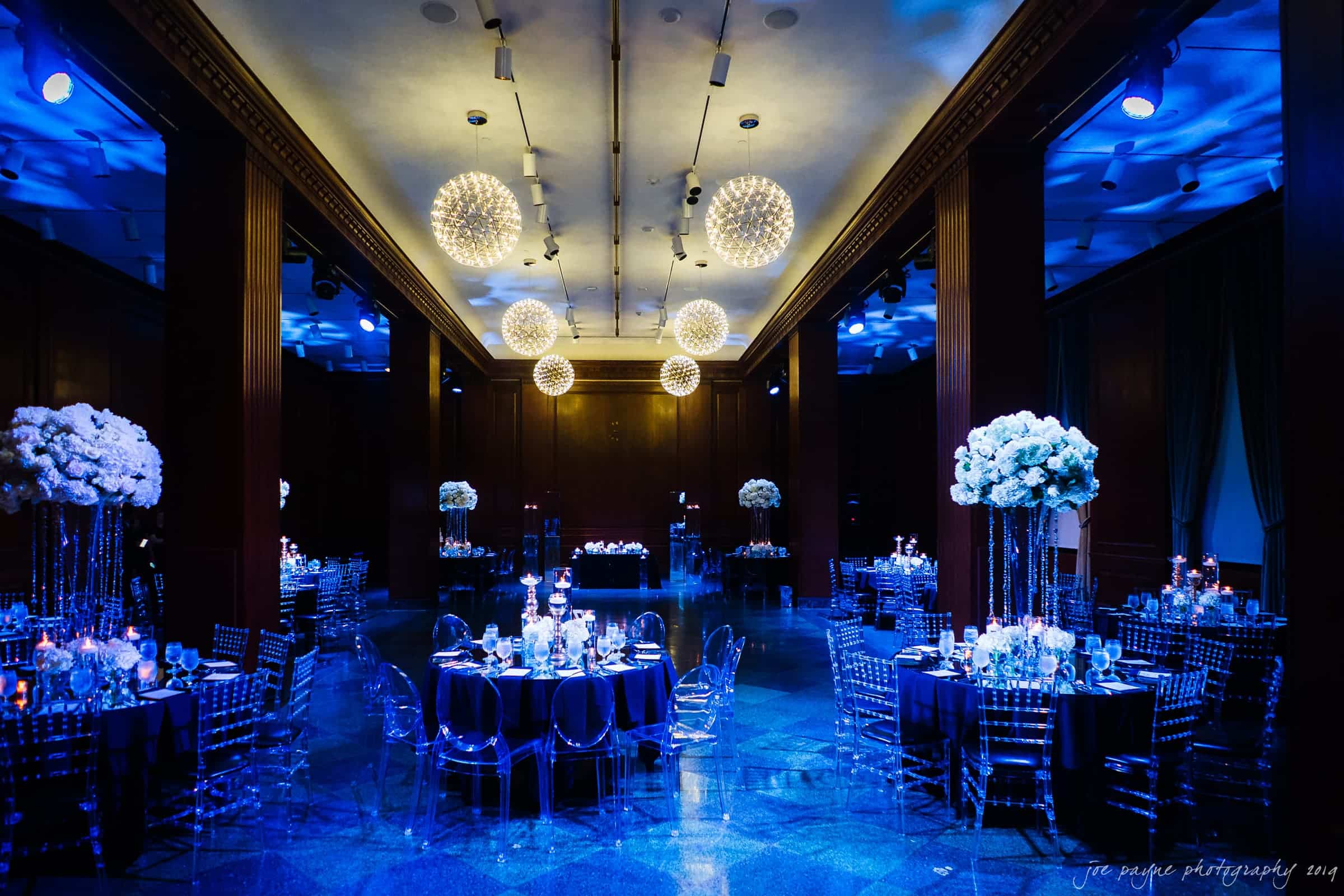 21c museum hotel durham wedding - kortne & terry Elegant banquet hall with blue lighting, featuring round tables with dark tablecloths and clear chairs. Tall floral centerpieces and glowing chandeliers create a luxurious atmosphere.
