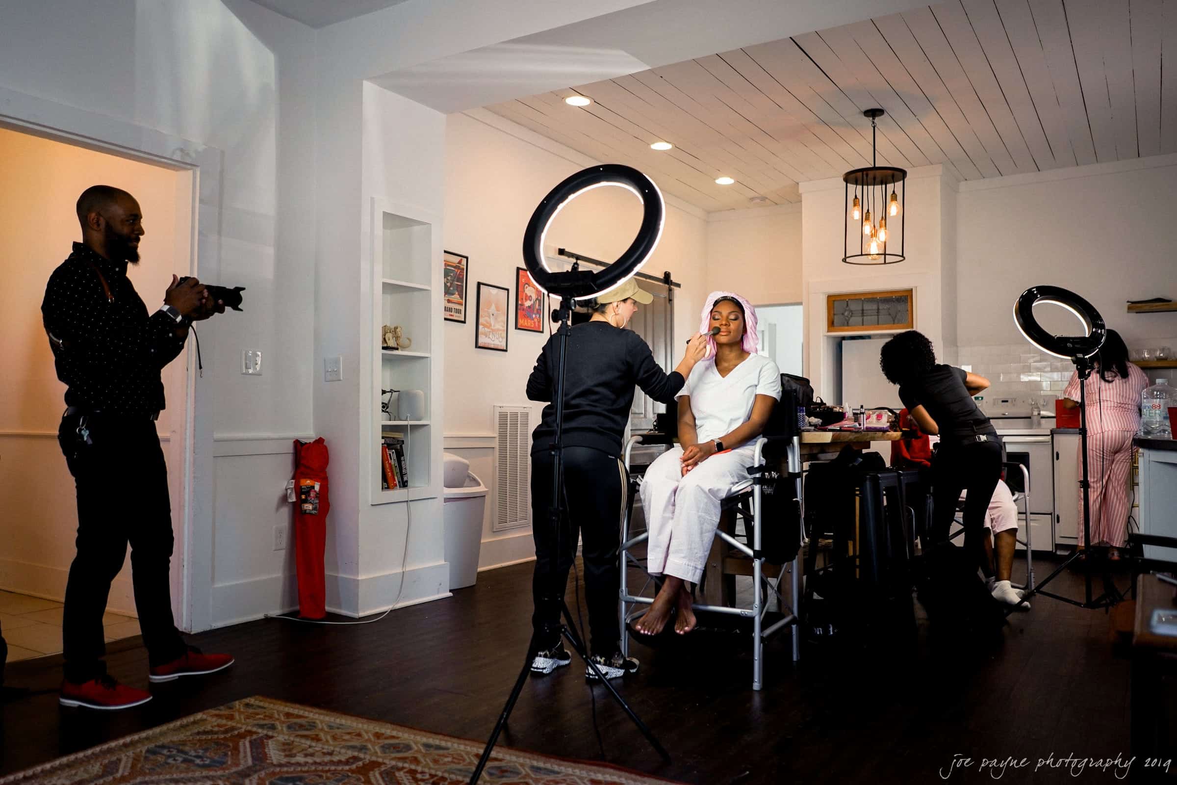melrose knitting mill wedding photography - johnique & derrick A make-up artist applies makeup to a seated woman under a ring light in a well-lit room. Two other people are present: one holds a camera, and another works at a table. The setting is relaxed and creative.