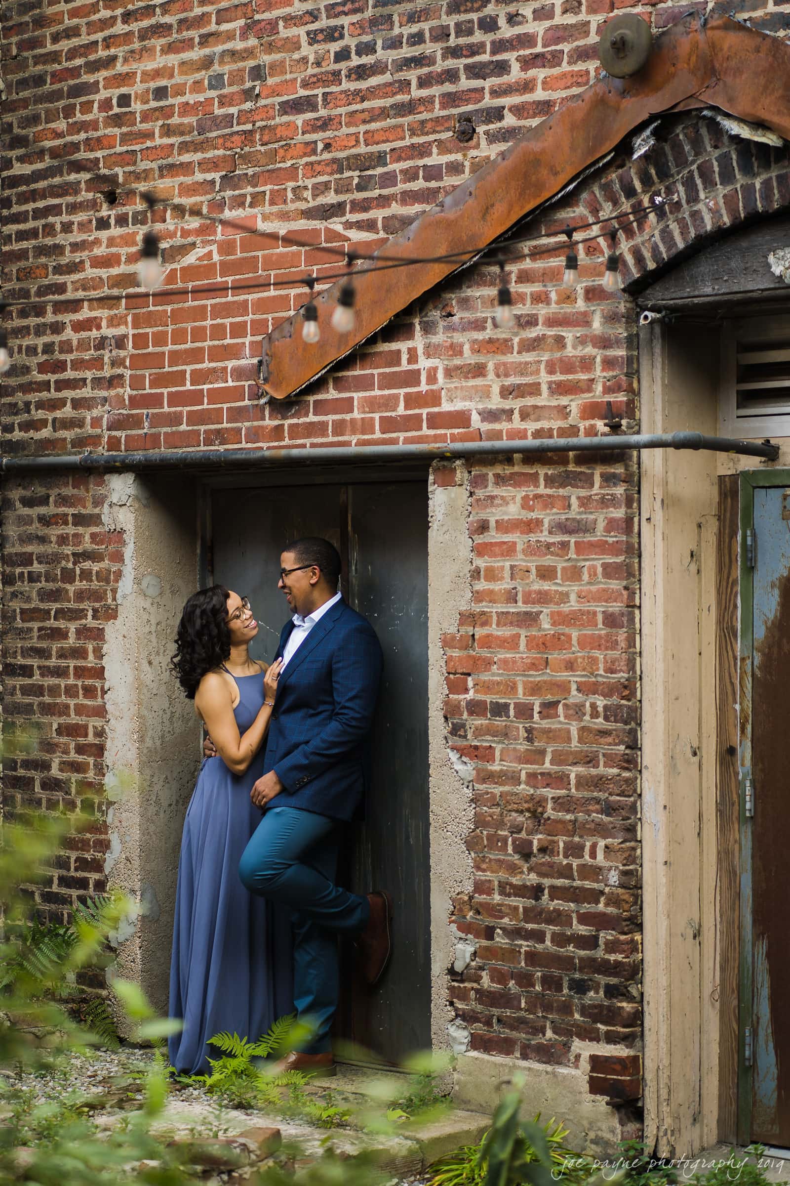 durham nc engagement photography | krystina & justin A couple stands close together, smiling at each other fondly. The woman is wearing a long blue dress, and the man is wearing a blue suit. They are leaning against a rustic brick wall adorned with string lights.