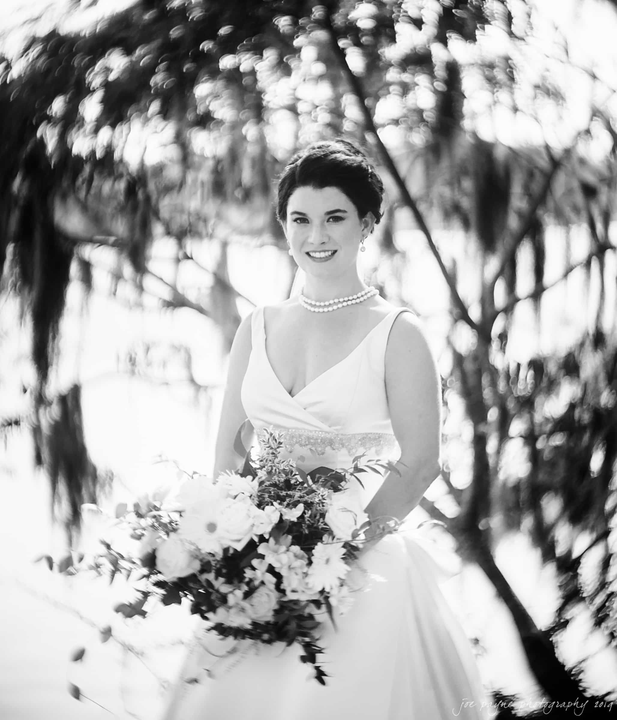 A bride in a white dress smiles while holding a bouquet. She stands outdoors, with blurred foliage in the background. The image is black and white, highlighting the elegance of her attire and features.