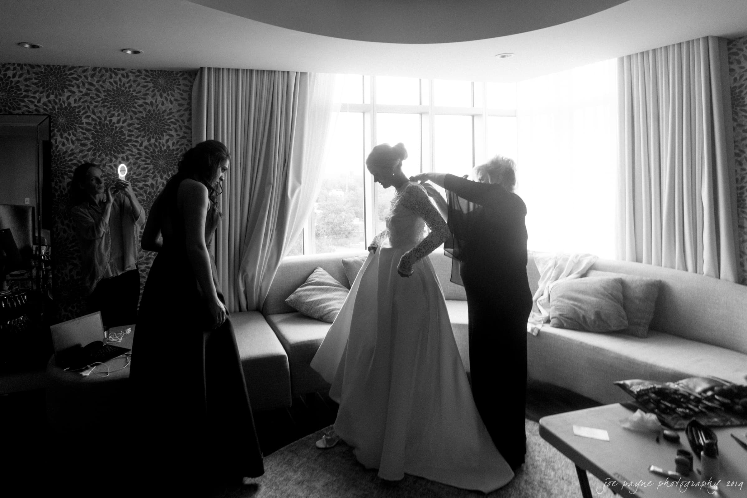 A black and white photo of a bride getting ready in a room with large windows. Three people assist her, adjusting her dress and hair. The atmosphere is calm and intimate, with soft light coming through the curtains.