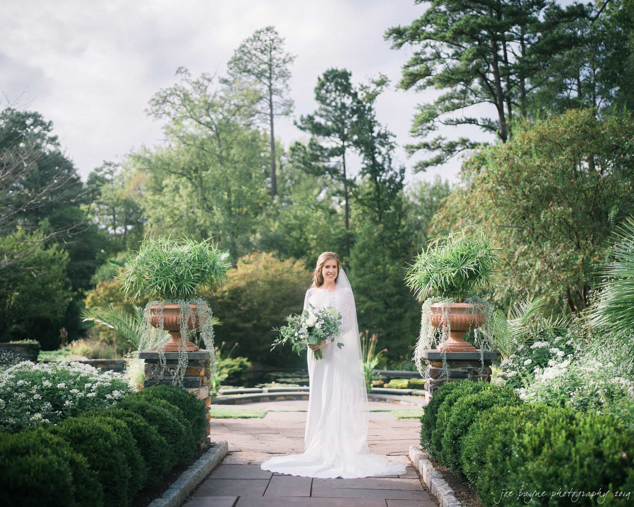 A bride in a long, flowing white dress stands outdoors on a stone pathway, surrounded by lush greenery and large potted plants. She holds a bouquet and smiles softly, with a garden and trees in the background under a slightly overcast sky.