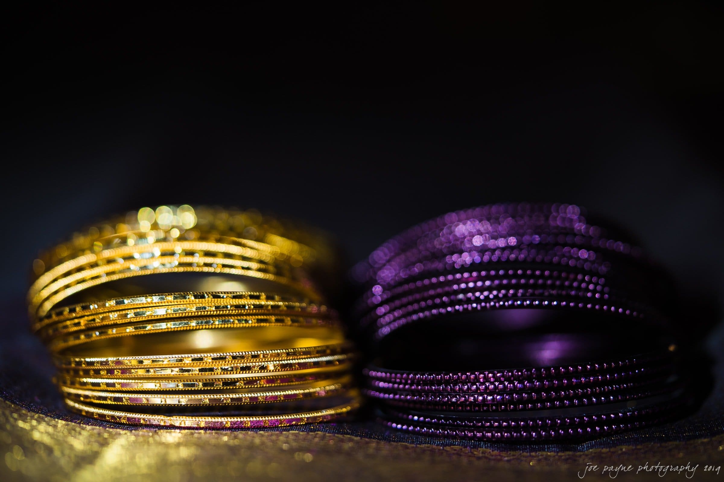 Close-up of several gold and purple bangles stacked together against a dark background, highlighting their intricate textures and shimmering surfaces.