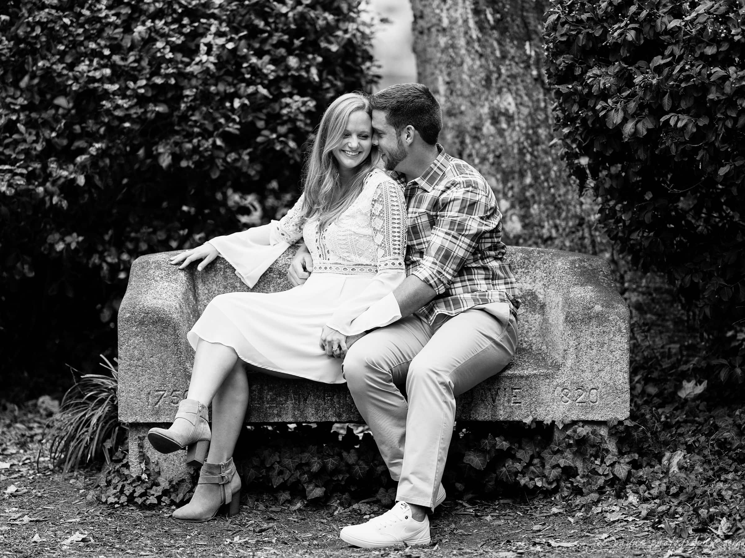 A couple sits closely on a stone bench in a park, surrounded by trees and foliage. The woman, wearing a white dress and ankle boots, smiles while the man, in a plaid shirt and light pants, leans in affectionately. The scene is captured in black and white.