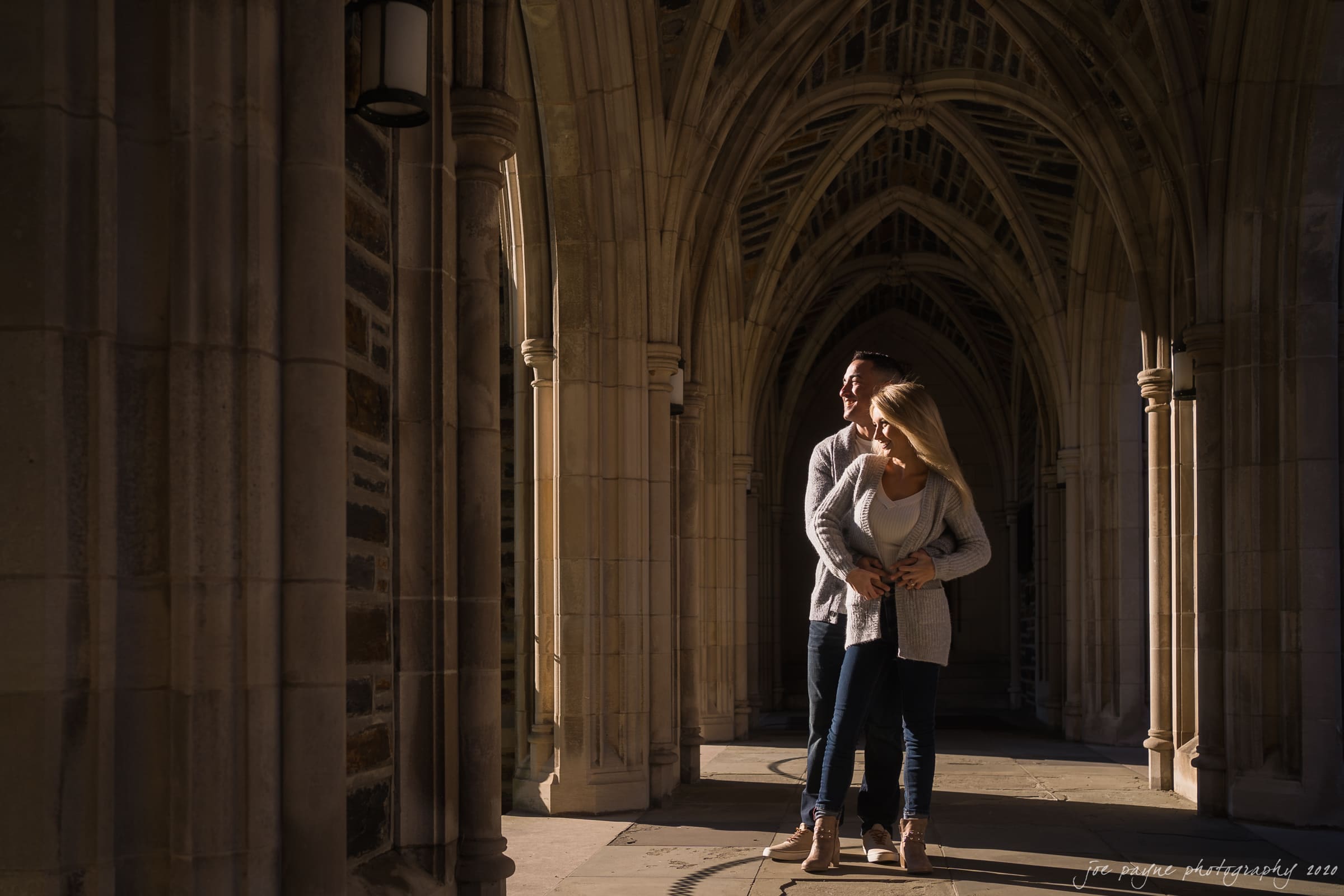 durham engagement photography - april & zach A couple stands in a sunlit, arched stone corridor. The woman is wearing a white top with a gray cardigan, and the man is wearing a gray sweater. They are embracing, with warm sunlight casting shadows on the columns and floor.