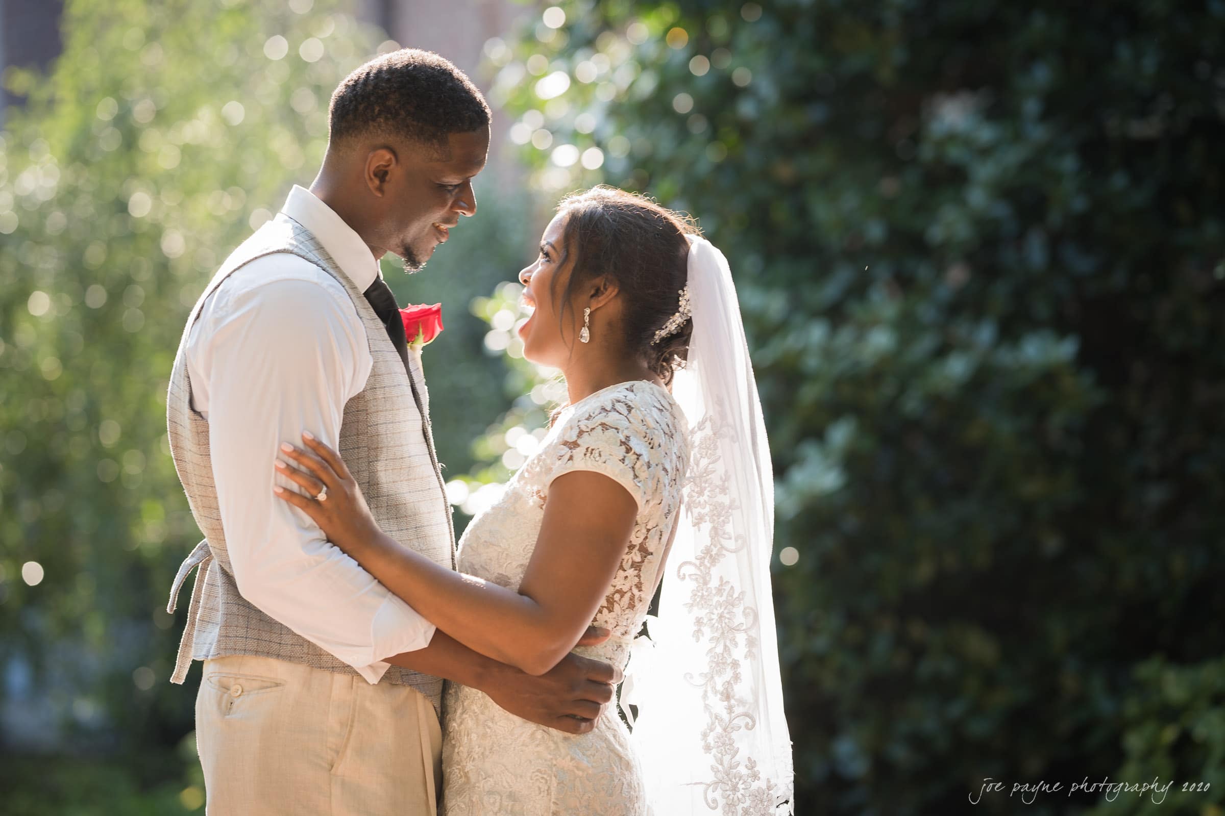 A bride and groom embrace outdoors in sunlight, surrounded by greenery. The bride wears a lace dress and veil, and the groom is in a white shirt and vest with a red rose boutonniere. They smile at each other in a serene, romantic setting.
