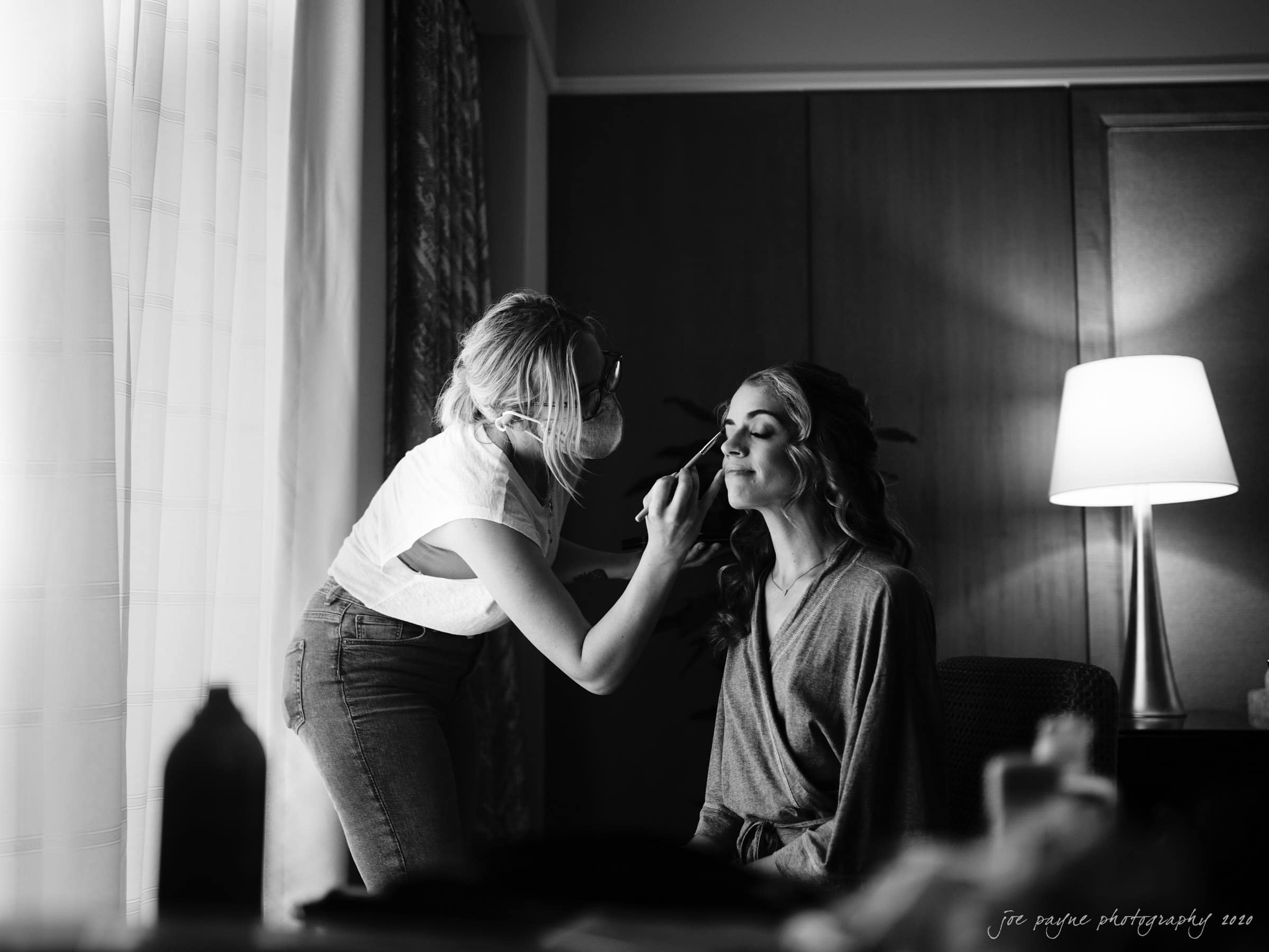 A black and white photo of a woman sitting in a chair, getting her makeup done by another person standing beside her. The setting appears to be a softly lit room with a lamp and a curtain in the background.